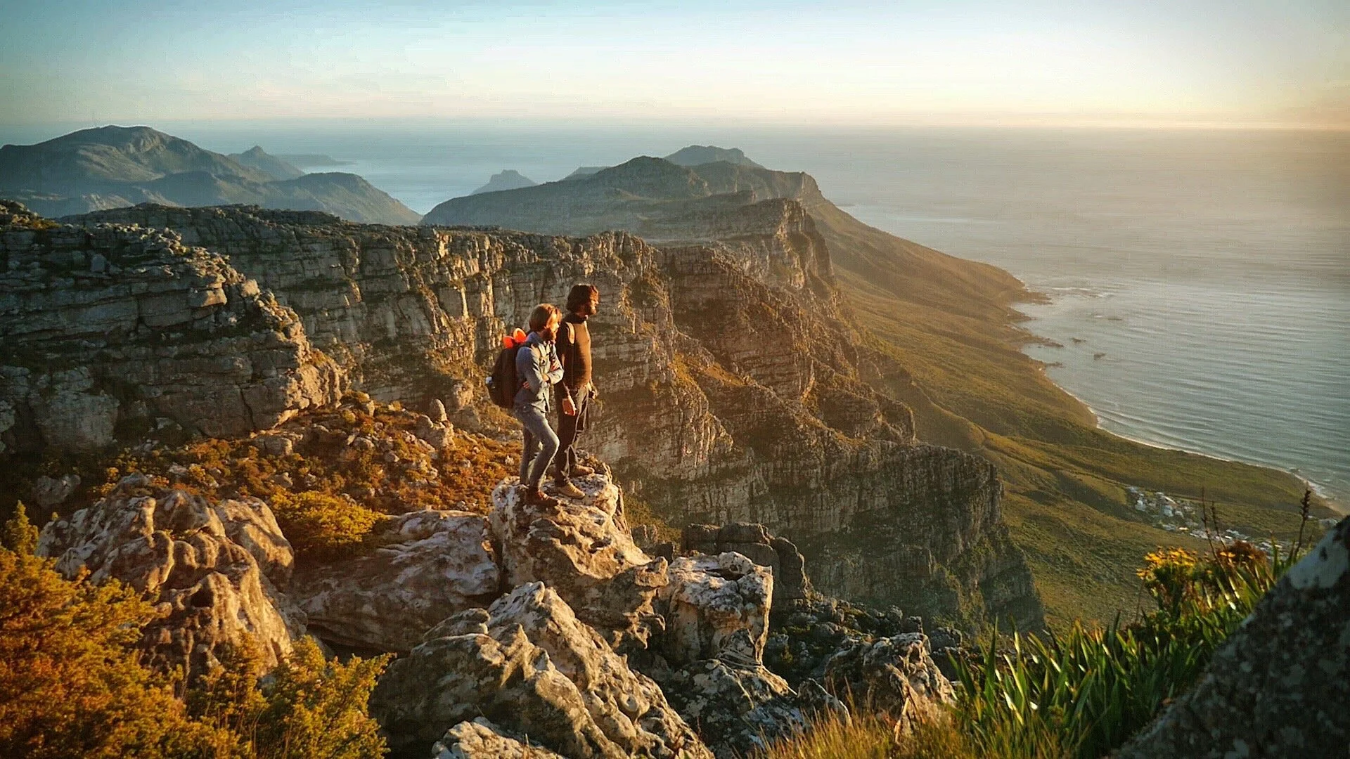 Two hikers standing on a rocky cliff overlooking a scenic coastline with rugged mountains and the ocean at sunset.