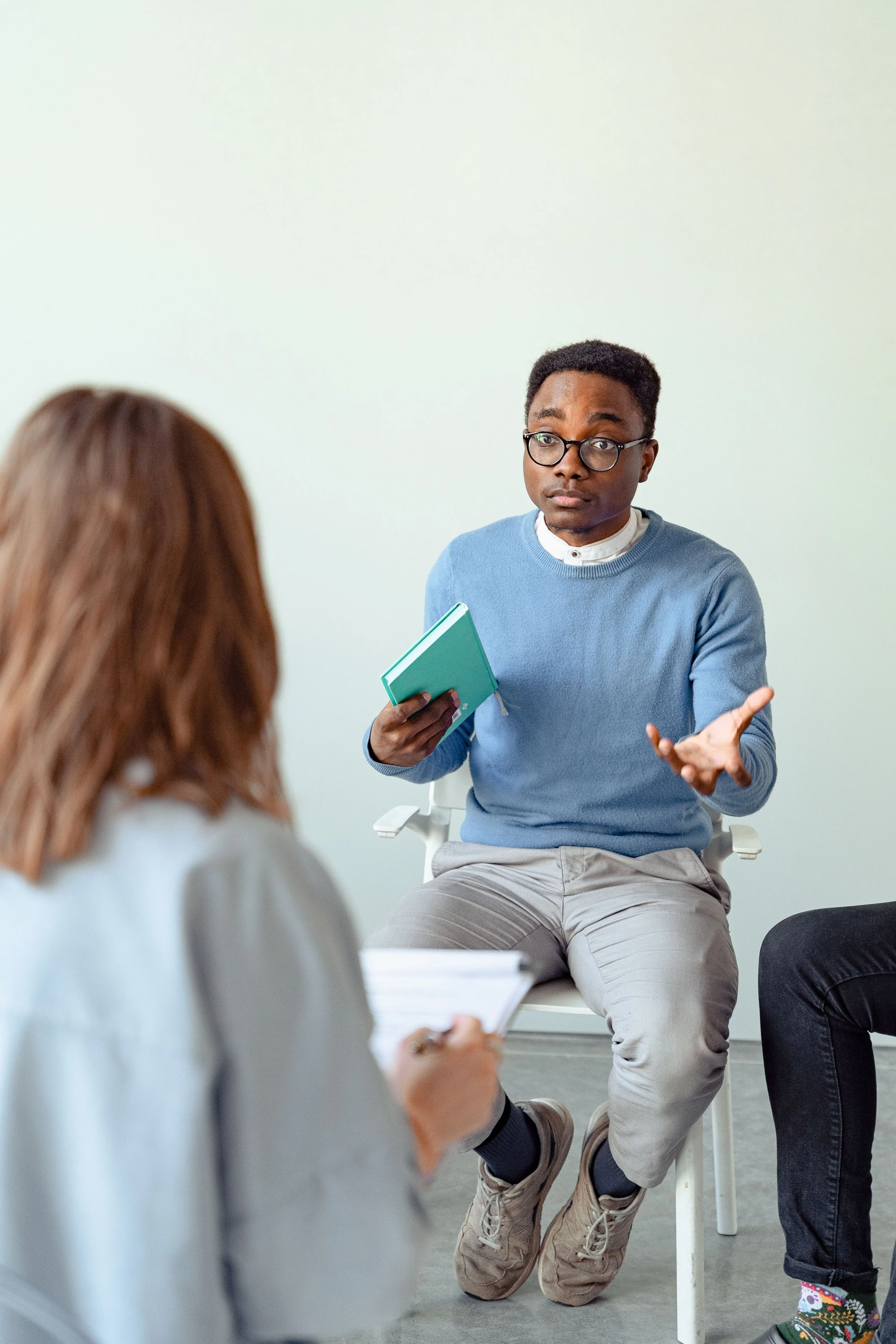 A man with glasses and a blue sweater sitting on a chair, talking to two women in a room with a plain cream wall. One woman has brown hair and is holding a notebook, the other woman has brown hair and is sitting on the floor, dressed casually.