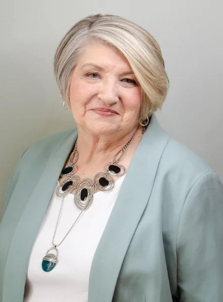 Portrait of an older woman with short blonde hair, smiling, wearing a light-colored blazer and a statement necklace with black and silver accents.