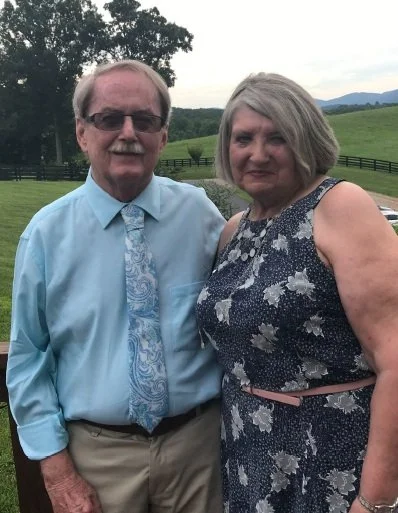 A man in glasses and a woman with short gray hair standing outdoors in a grassy field with trees and hills in the background.