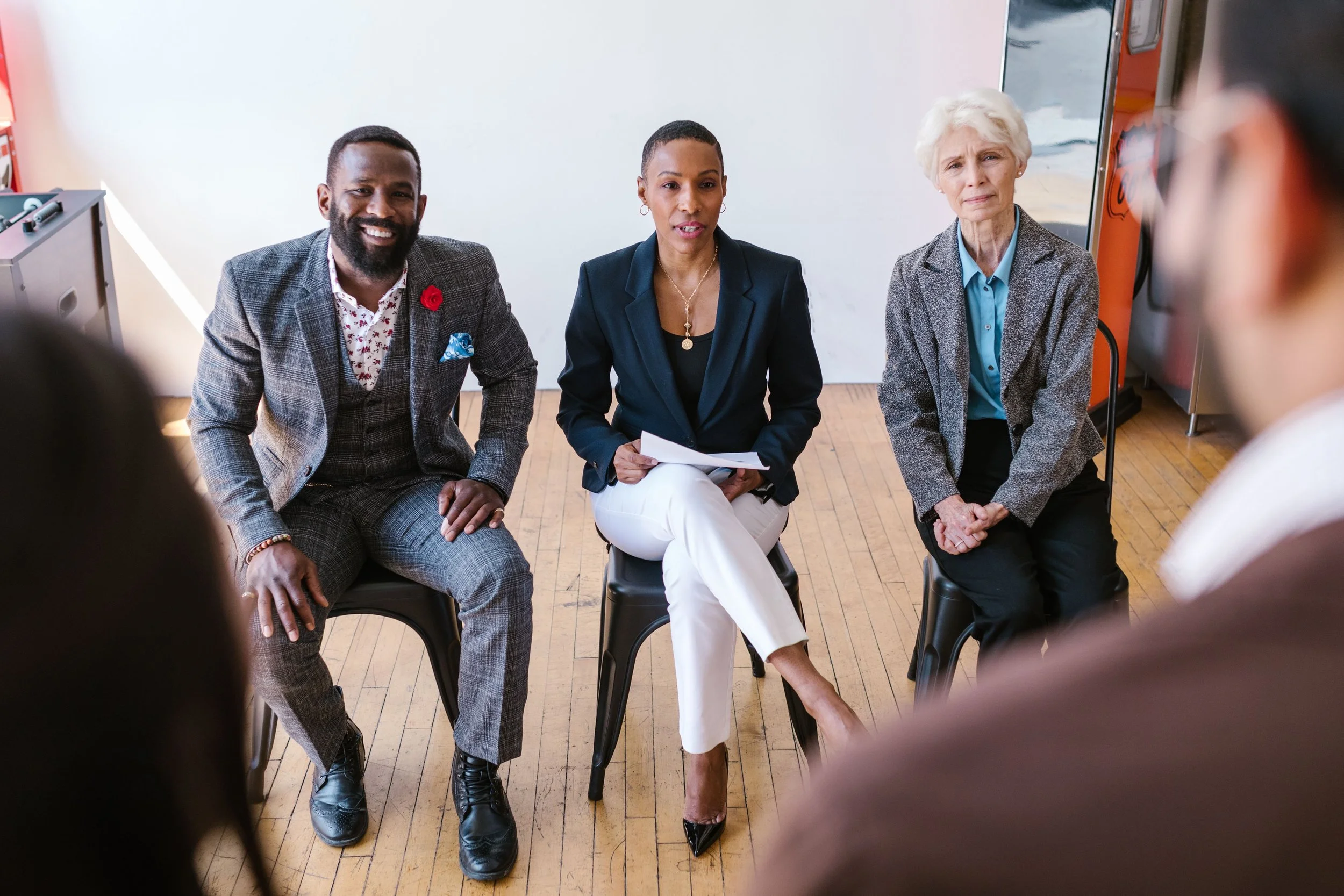 Three people sitting in chairs during a discussion or interview. The person on the left is a man with a beard, wearing a plaid suit jacket with a red flower pin and floral shirt. The person in the middle is a woman with short hair, wearing a black blazer and holding papers. The person on the right is an elderly woman with white hair, wearing a gray blazer and a blue shirt. The image appears to be taken in an indoor setting with wooden flooring.