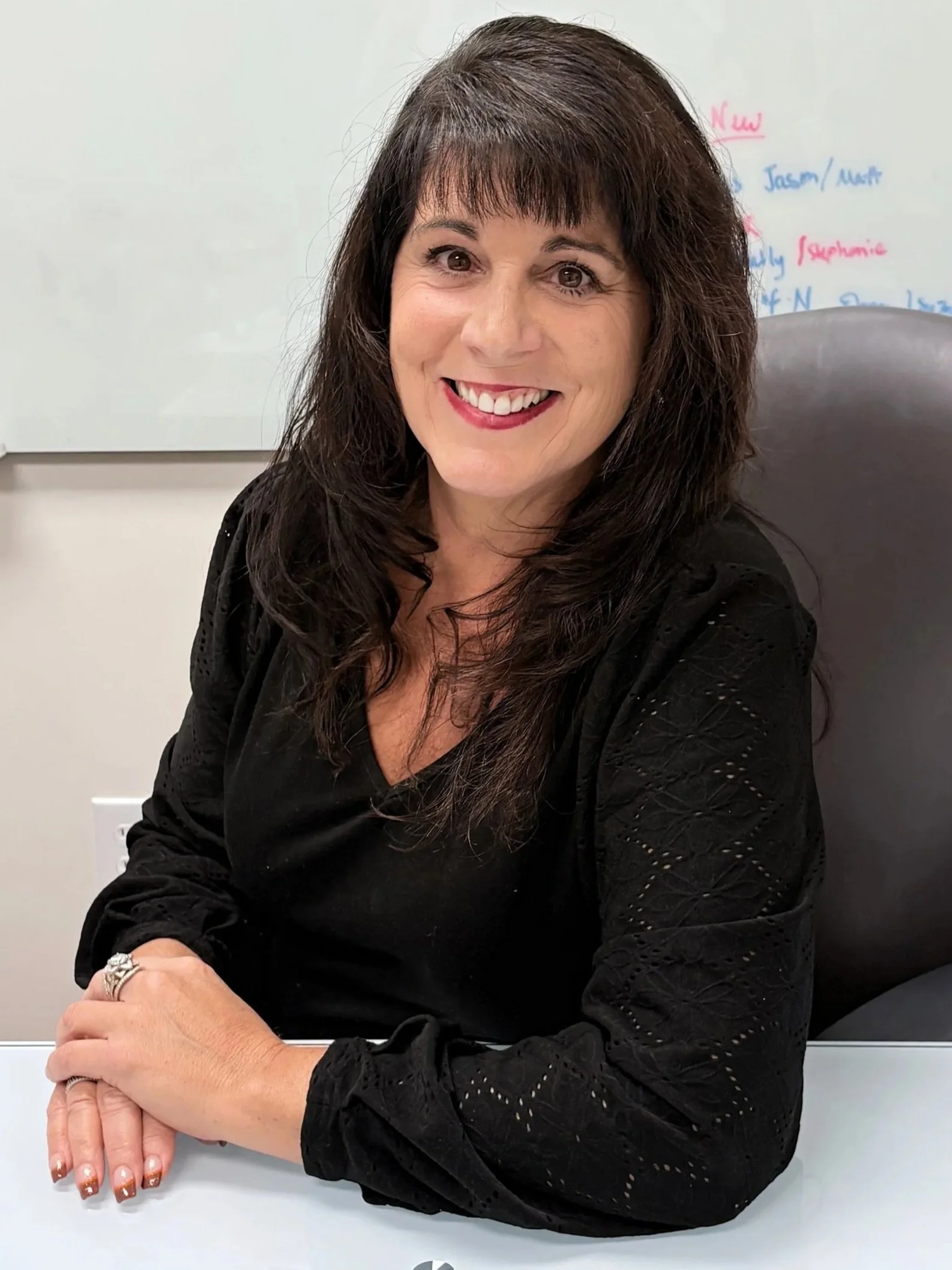 A woman with dark brown hair smiling, sitting at a desk with her arms folded on the surface, wearing a black blouse with eyelet details, in a room with a whiteboard in the background.