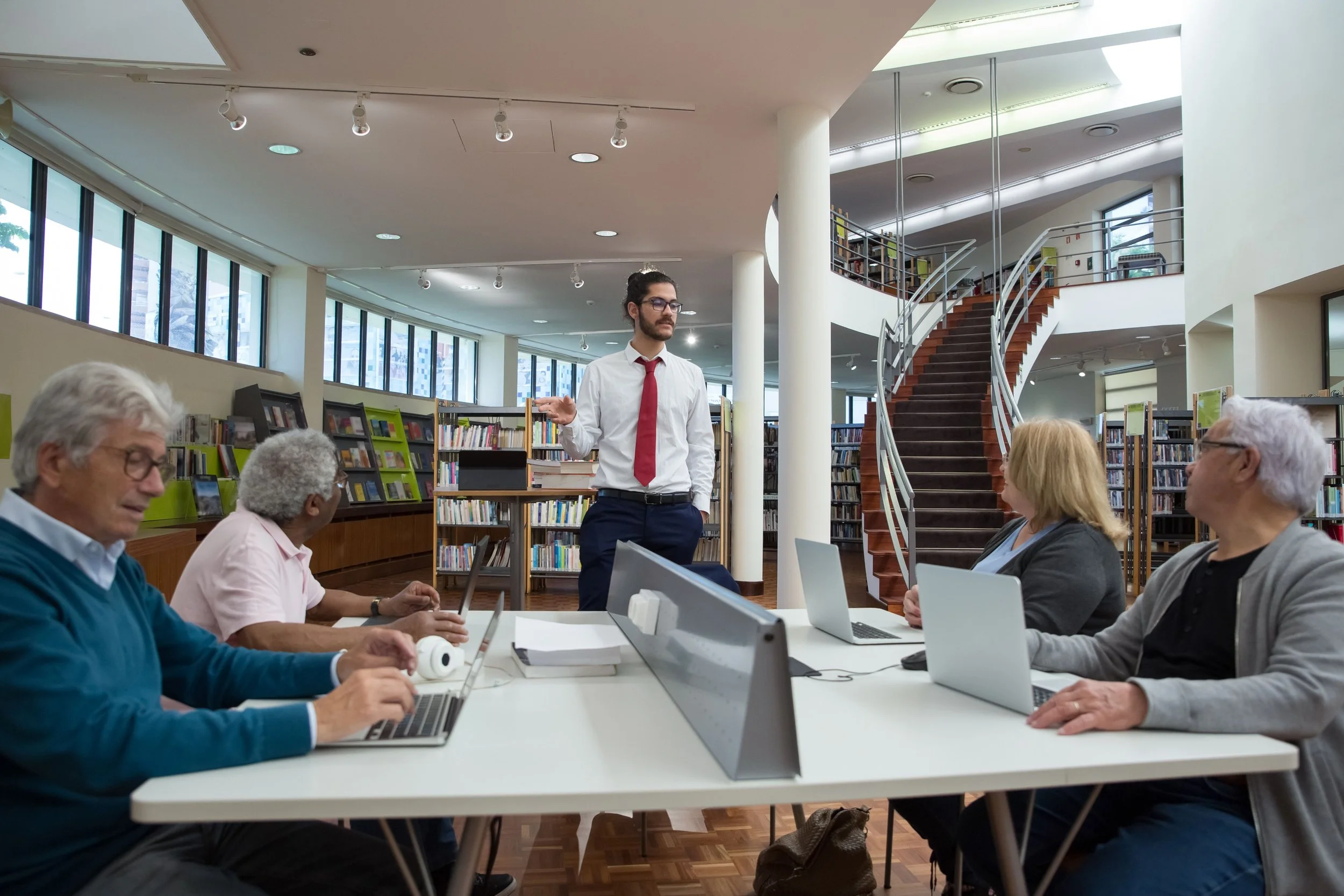 A man in a white shirt and red tie stands in front of a group of seated people with laptops in a modern library or conference room.