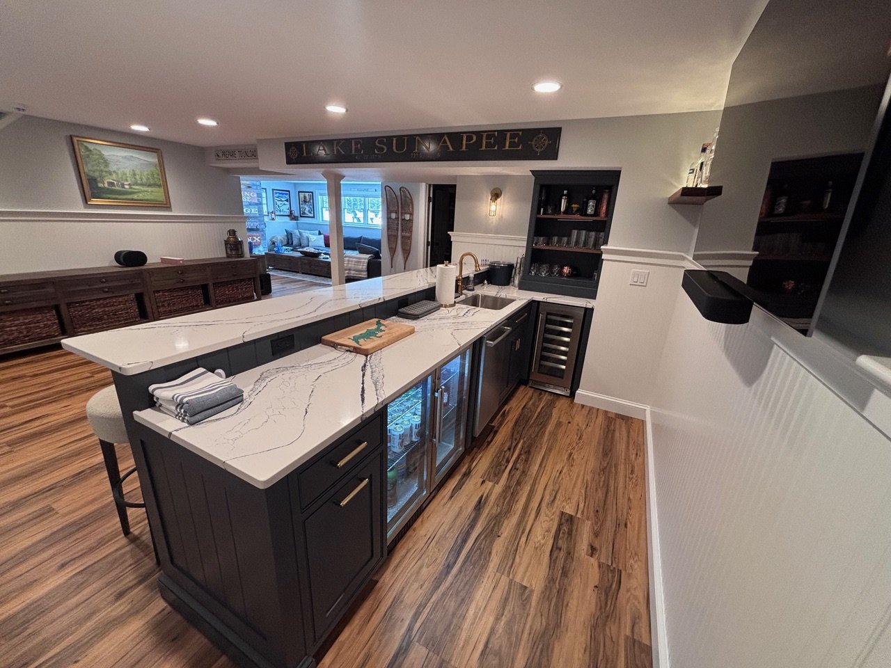 Modern kitchen with dark cabinetry, marble countertop, and a wine fridge under the counter. Open living space with a view of a sitting area and decorative sleds. Recessed ceiling lights and wood flooring throughout.