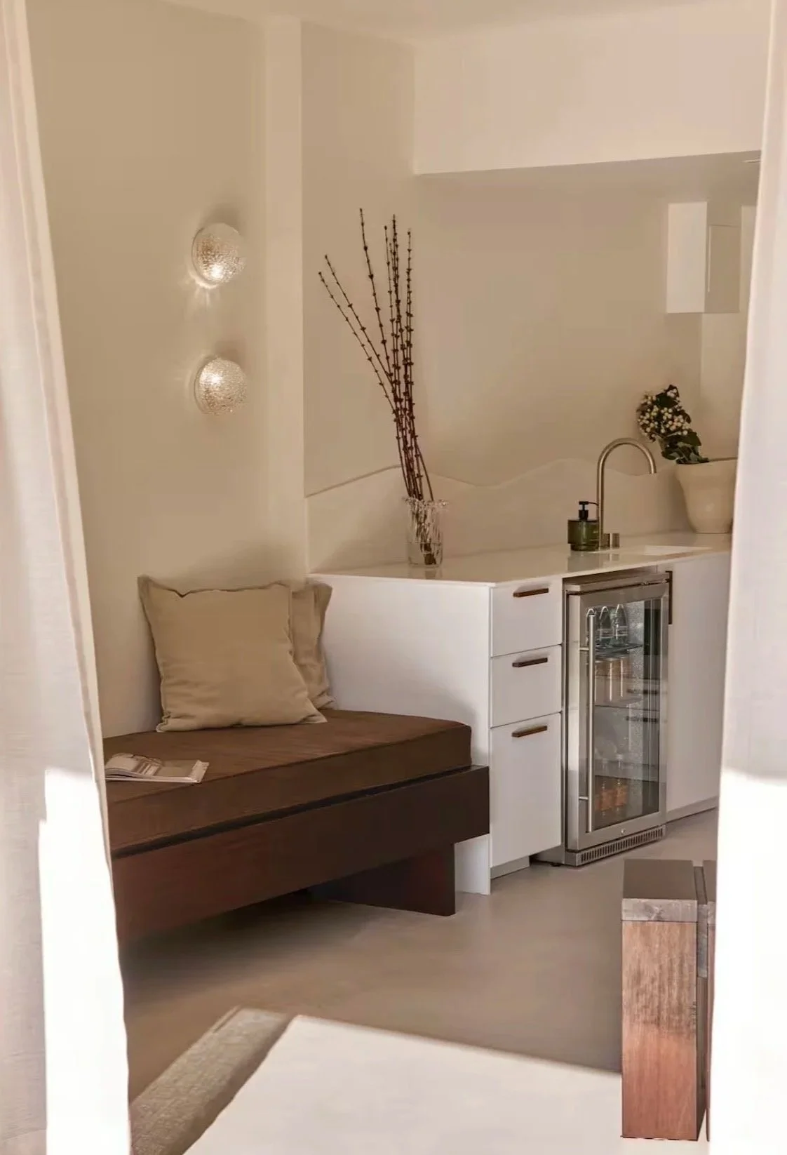A cozy corner with a wooden bench with a beige pillow, a white cabinet with a curved top, a small wine fridge, and decorative items like a plant in a white pot and tall dried branches in a vase, against a off-white wall with two textured wall lights.