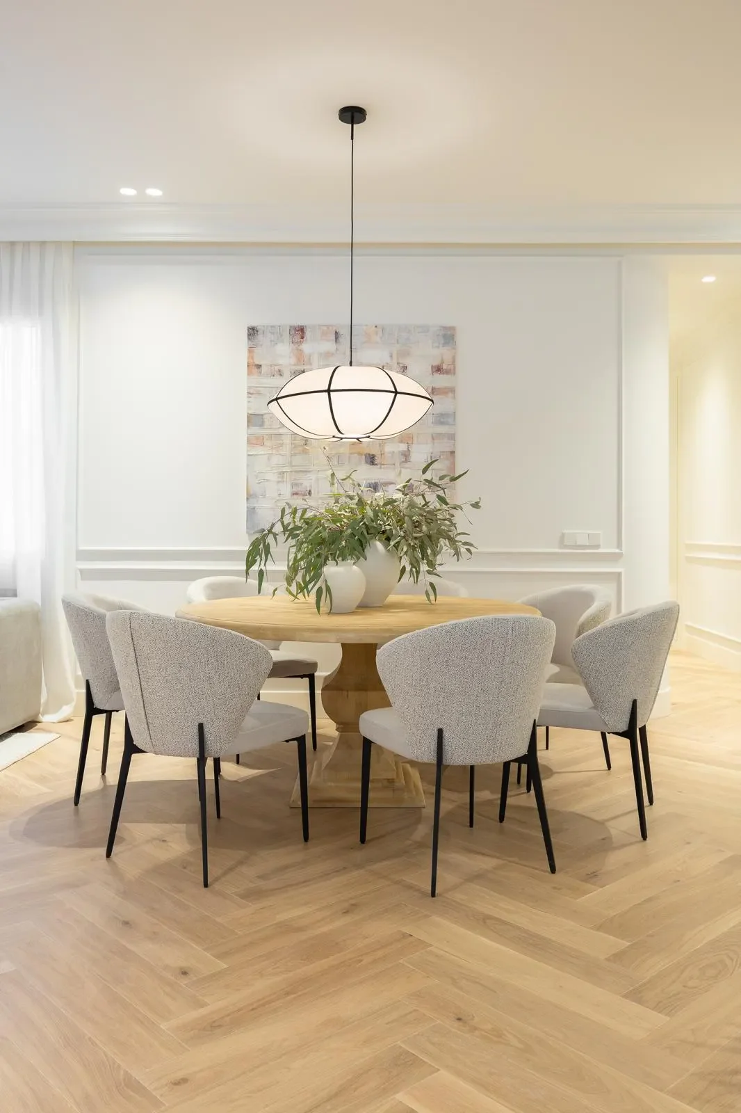 Dining room with a round wooden table, eight upholstered chairs, a hanging pendant light, and a large potted plant as decor.
