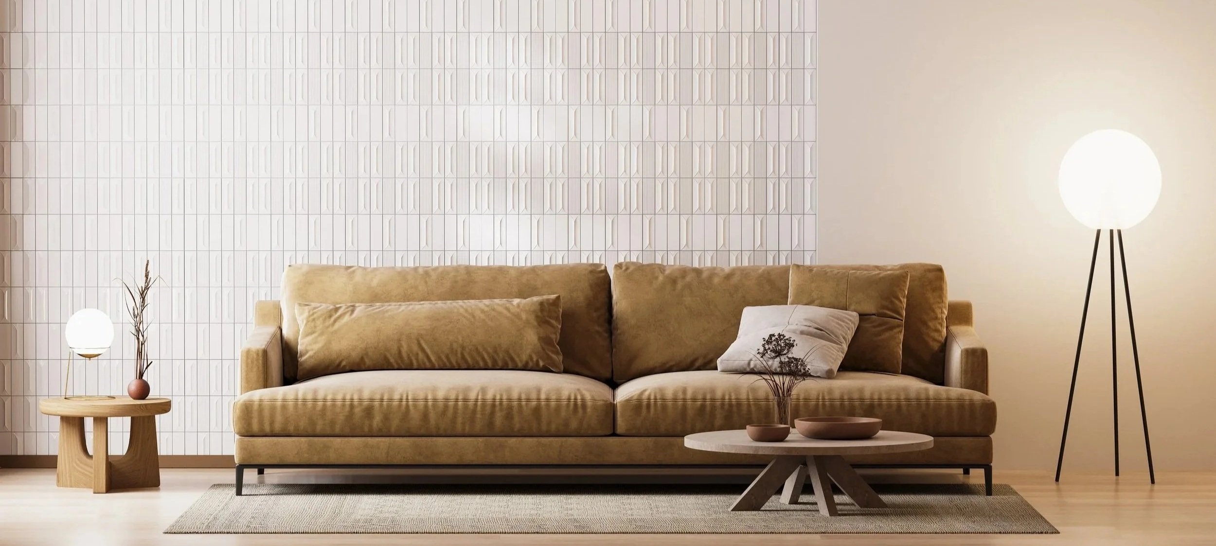 A living room with a tan velvet sofa, beige pillows, a wooden side table with a small round vase and dried plant, a round coffee table with decorative bowls and dried flowers, a textured white wall, a tall tripod floor lamp with a glowing white globe, and a large area rug.