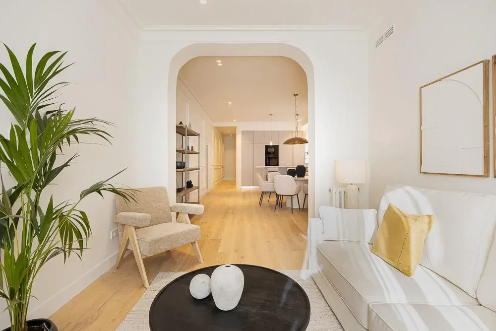 Living room with white walls, beige and white furniture, wooden flooring, and decorative wall art. Includes a potted plant, armchair, white sofa with a pillow, side lamp, and black coffee table with white vases. Opens into dining and kitchen area.