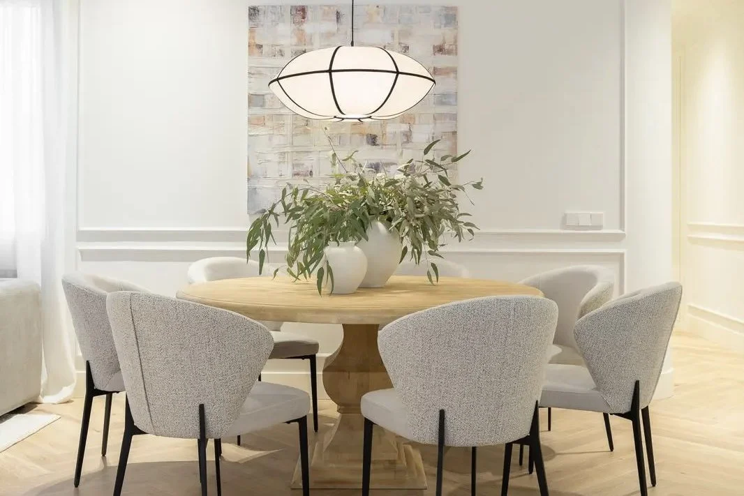Dining room with a round wooden table, six upholstered chairs, a white vase with green foliage on the table, and a modern pendant light overhead.