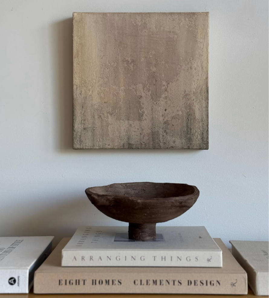 A minimalist interior decor scene featuring a square abstract painting on the wall, a brown ceramic bowl on two books, and additional books on either side.