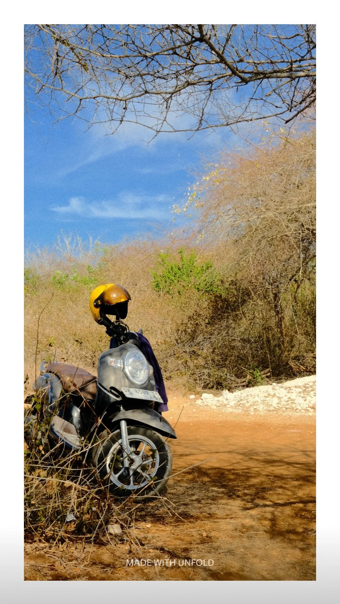 A black and brown scooter with a yellow helmet resting on its handlebar parked on a dirt road surrounded by dry, leafless bushes and trees under a blue sky.