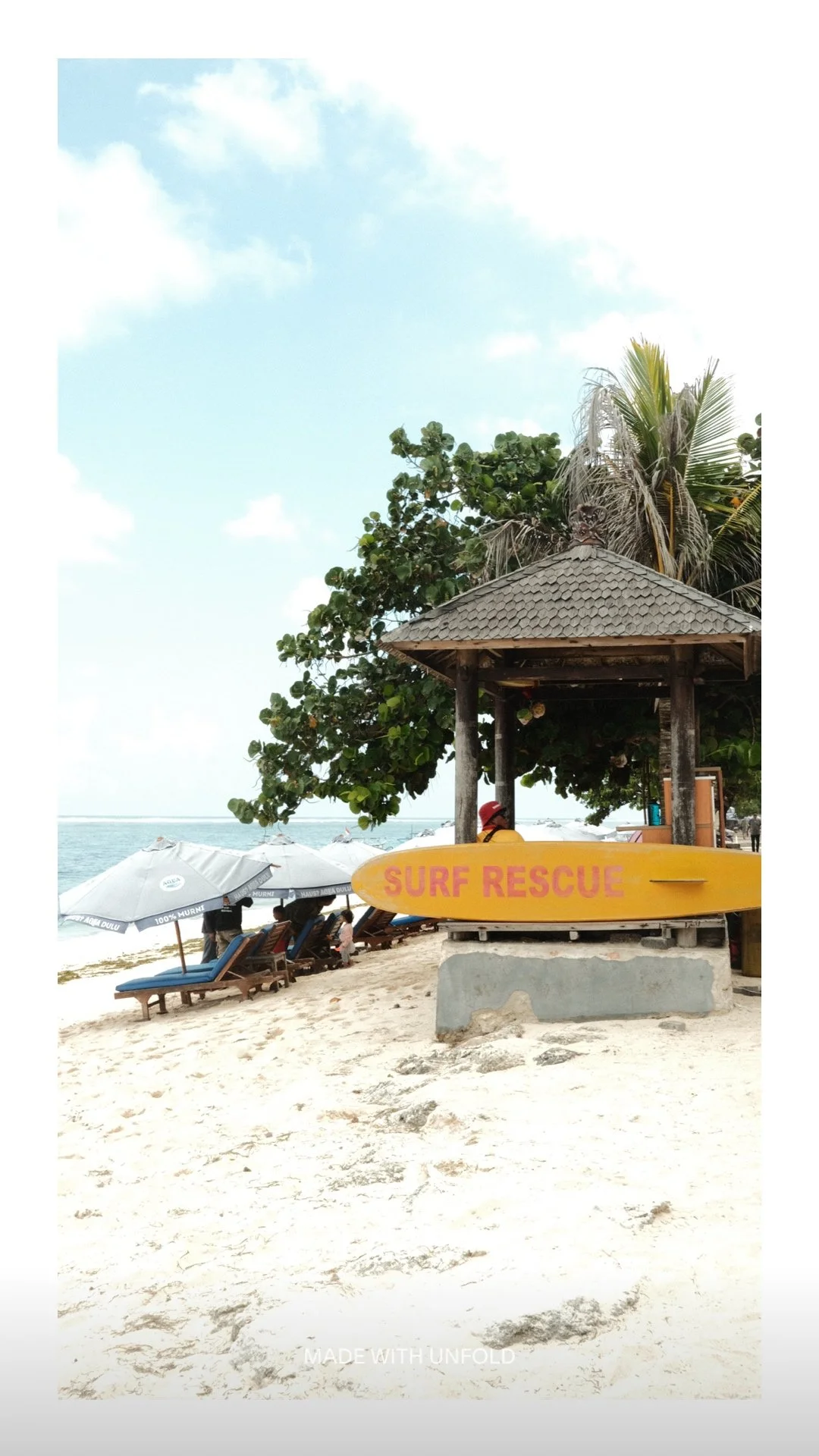 A beach scene with lounge chairs under umbrellas, a wooden hut with a yellow surf rescue board in front, palm trees, and the ocean in the background.