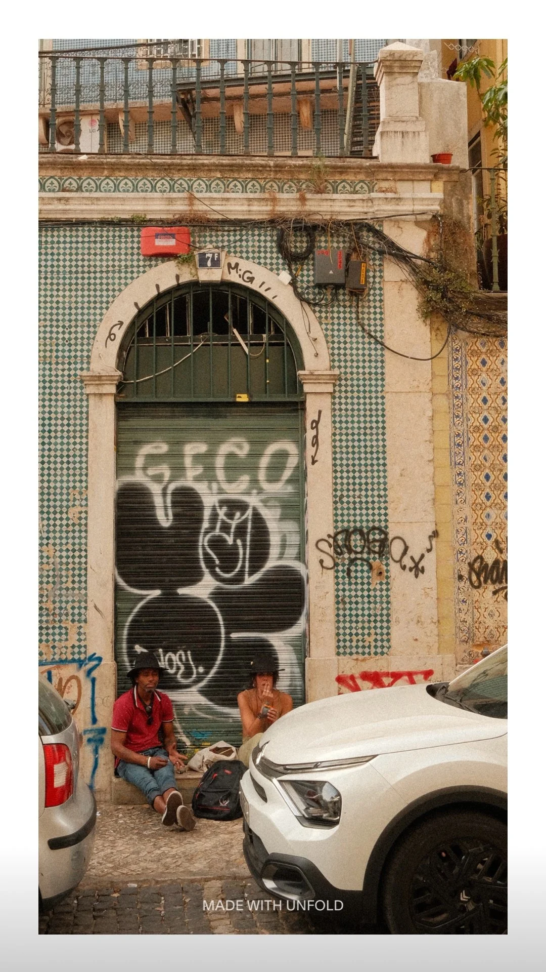 People sitting on the sidewalk in front of a graffiti-covered metal shutter, with parked cars nearby and an old building with decorative tiles behind them.