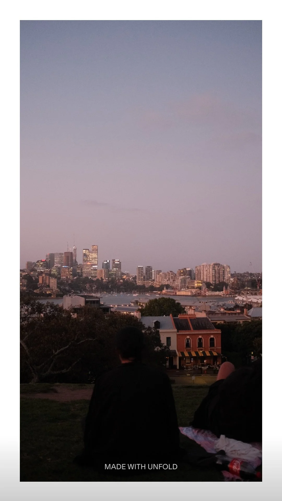 View of a city skyline at dusk with tall buildings and a river in the foreground, seen from a park with two people sitting on the grass.
