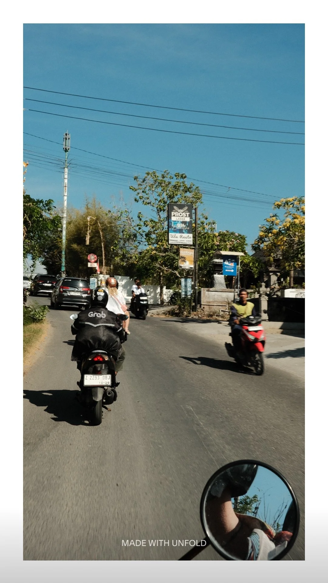 Street scene with motorcycles, cars, and people riding scooters, with trees, buildings, and a billboard in the background under a clear blue sky.