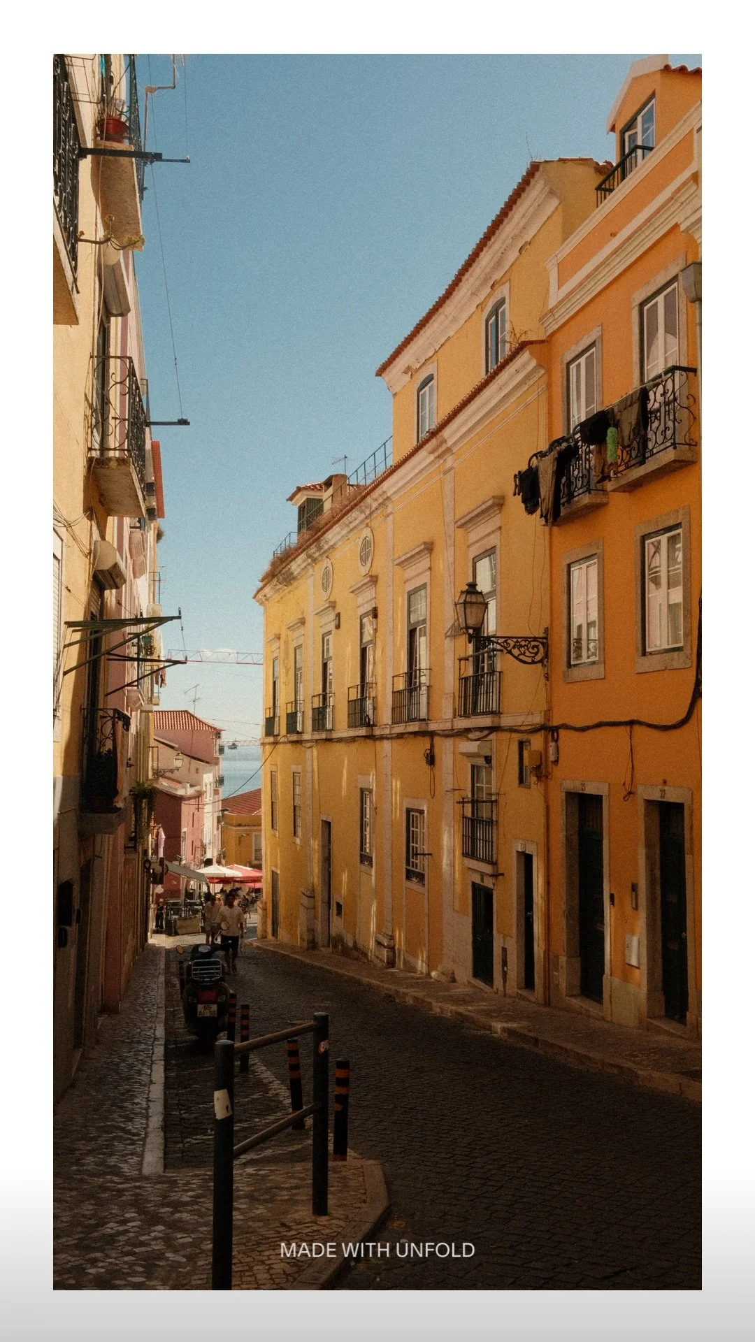 A narrow cobblestone street lined with yellow and pink apartment buildings under a clear blue sky, with a few pedestrians and parked bicycles.