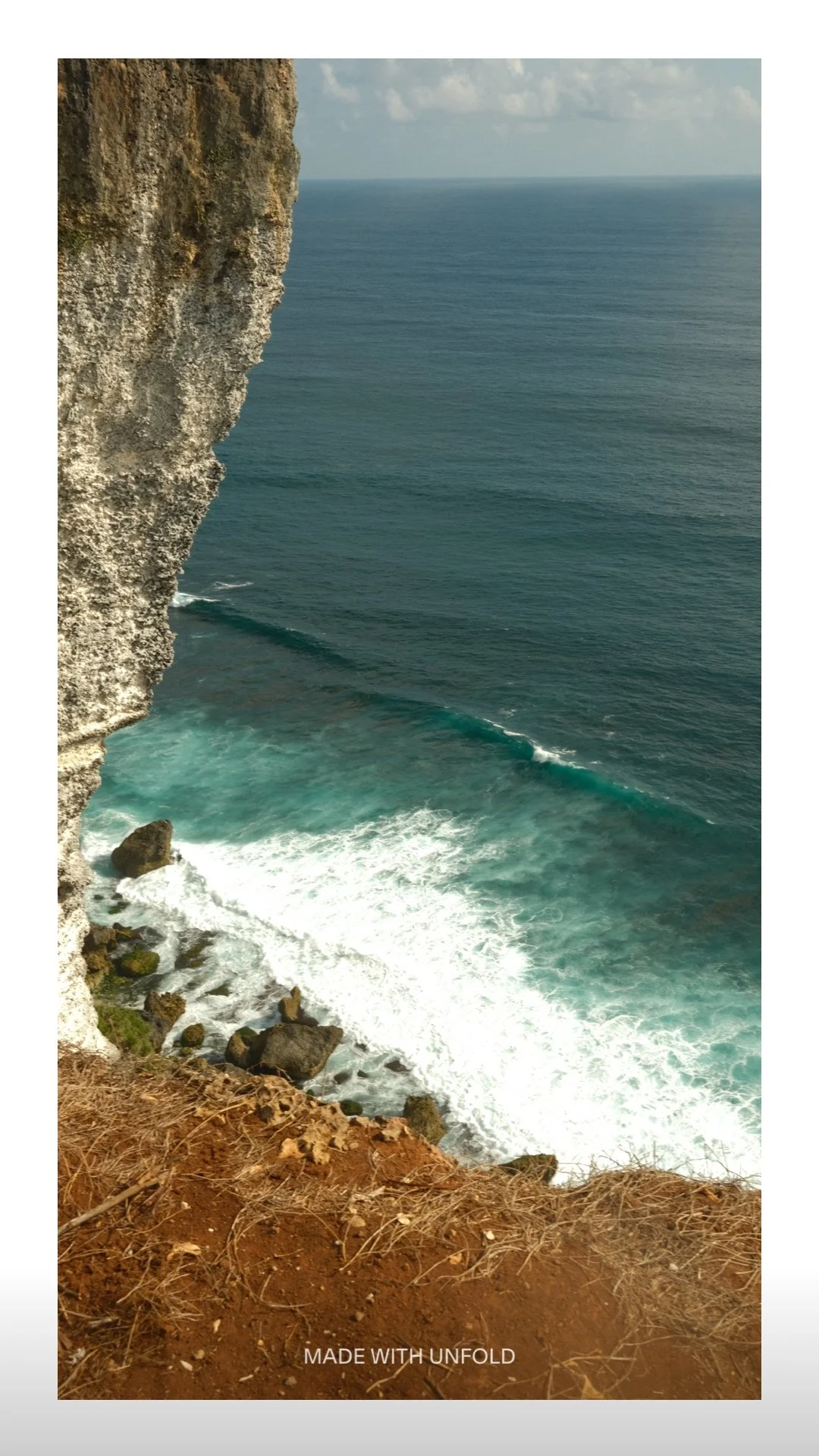 Cliffside view of the ocean with waves breaking against rocks at the base and a cloudy sky in the background.