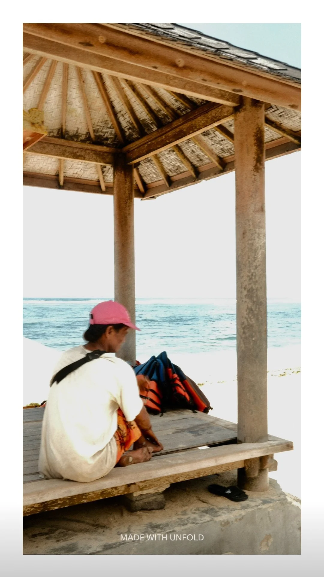 A person wearing a pink cap and white shirt sitting on a wooden platform under a thatched roof, overlooking the ocean with a backpack beside them.