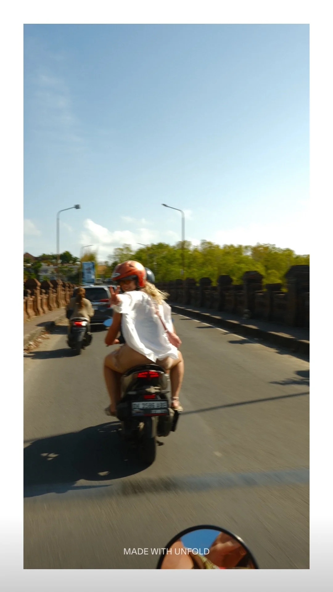 A woman and a child riding a motorcycle on a sunny street, waving at the camera, with another person riding a scooter in front of them and trees and streetlights in the background.