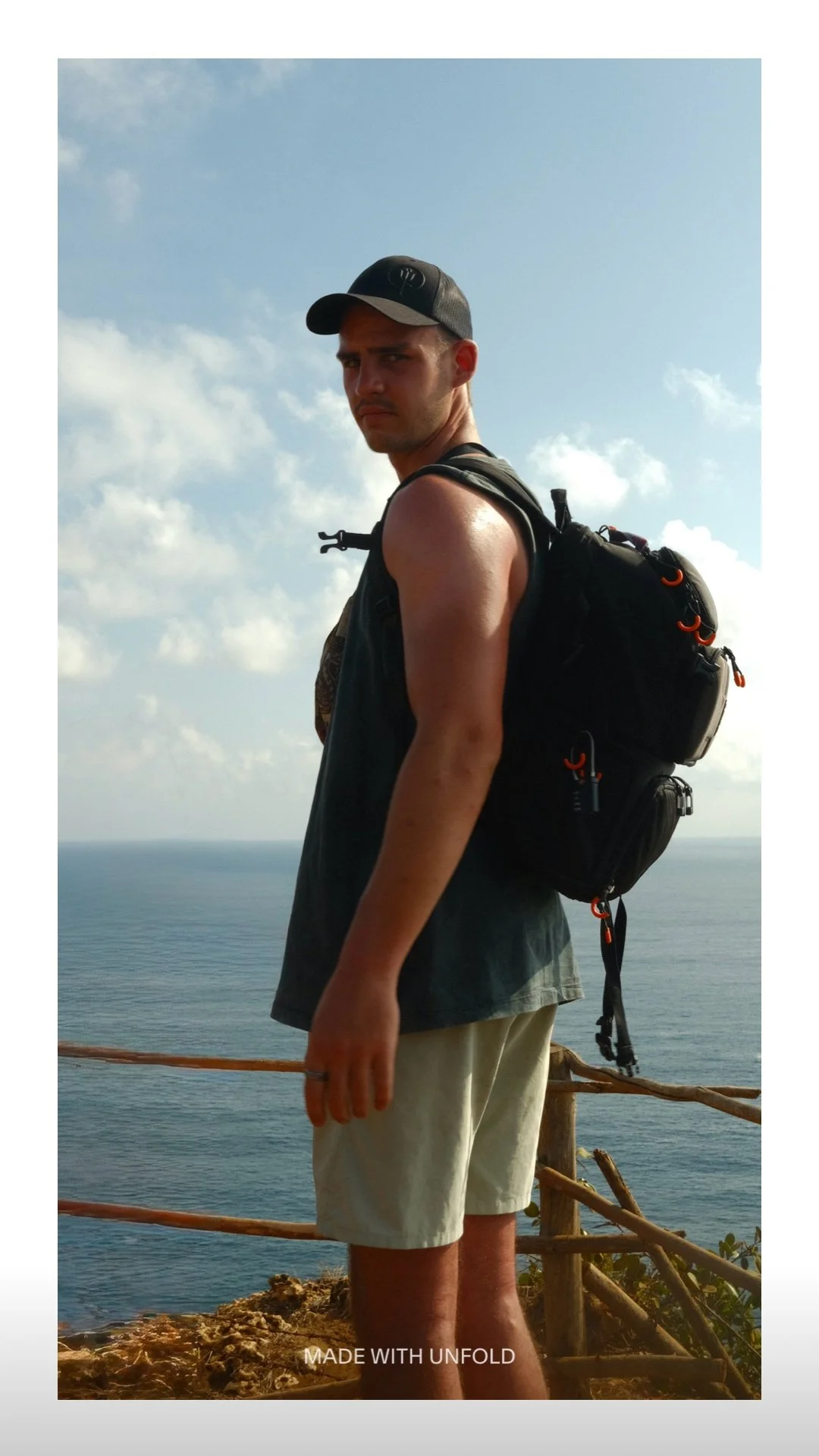 Young man with a backpack standing on a cliff overlooking the ocean, wearing a black cap, sleeveless shirt, and beige shorts, with a wooden railing behind him and cloudy sky above.