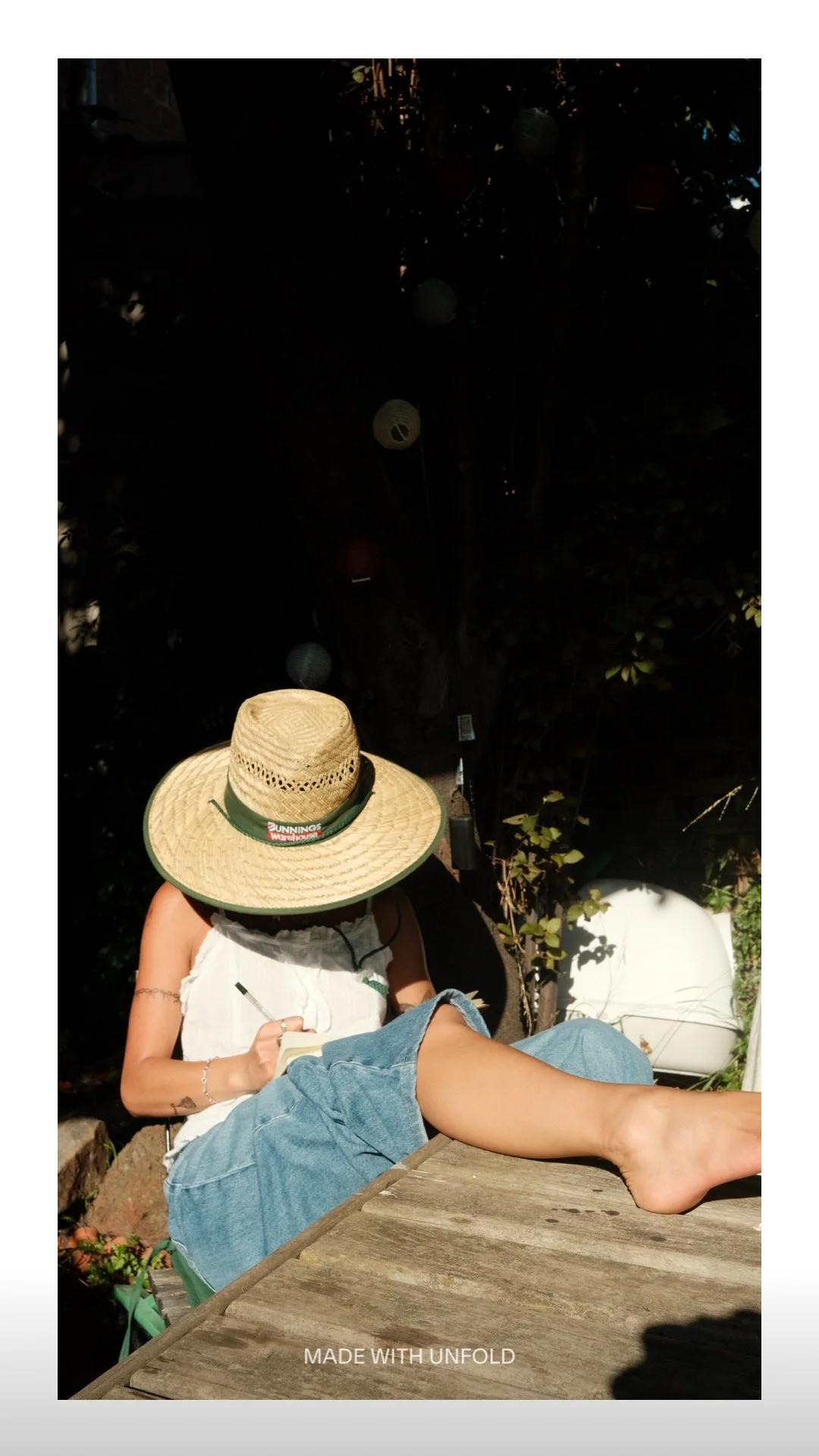 A person wearing a large straw hat with a green band sitting outdoors and writing in a notebook. They are sitting on a wooden surface with one leg extended and barefoot.