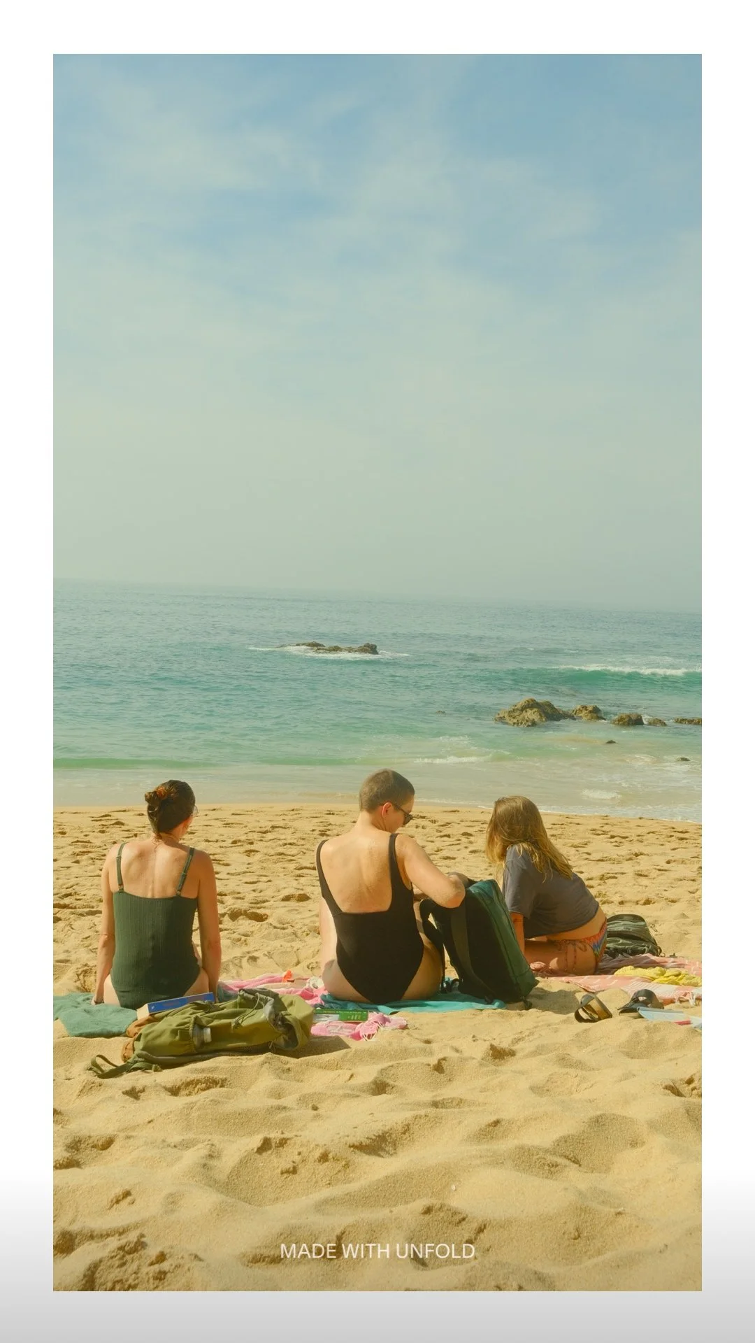 Three women sitting on a beach towel facing the ocean, with rocks in the water and a clear sky above.