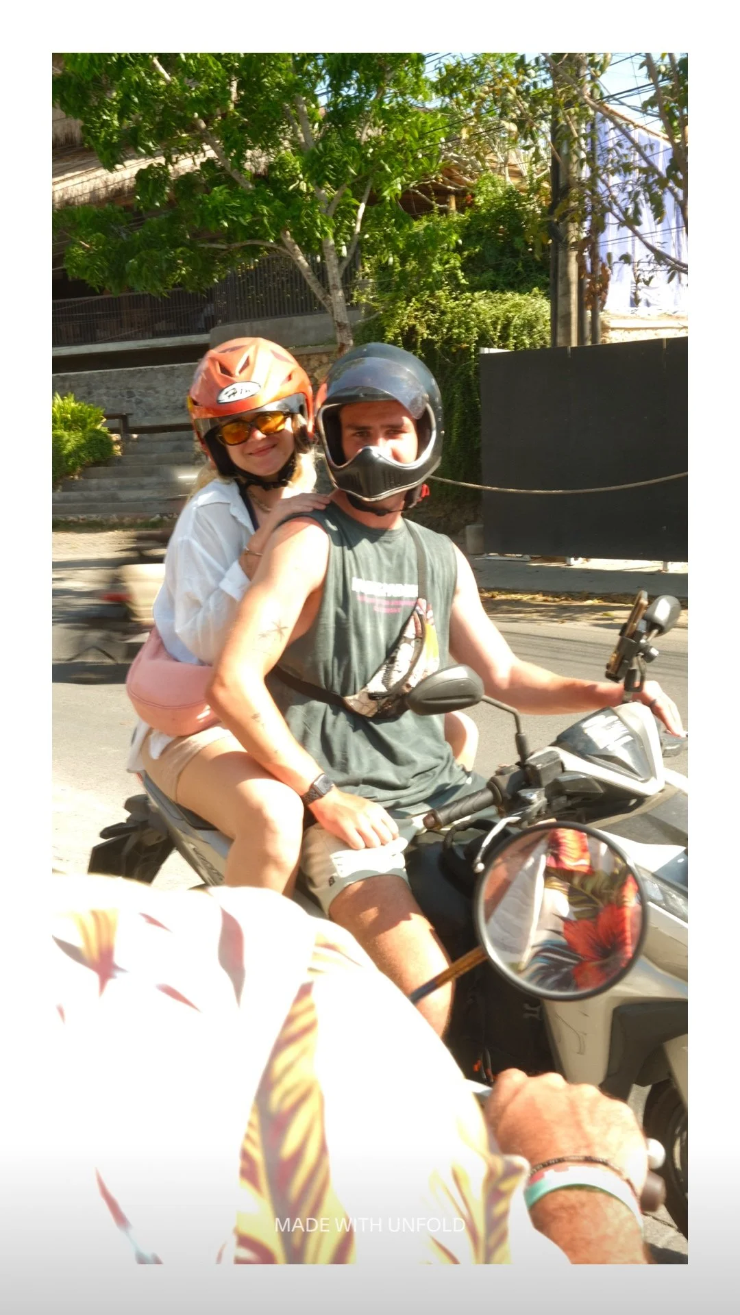 Two people on a motorcycle, the person in front wearing a helmet and sleeveless shirt, the passenger in back wearing sunglasses, a helmet, a white shirt, and light-colored shorts, riding on a sunny street with trees and a building in the background.