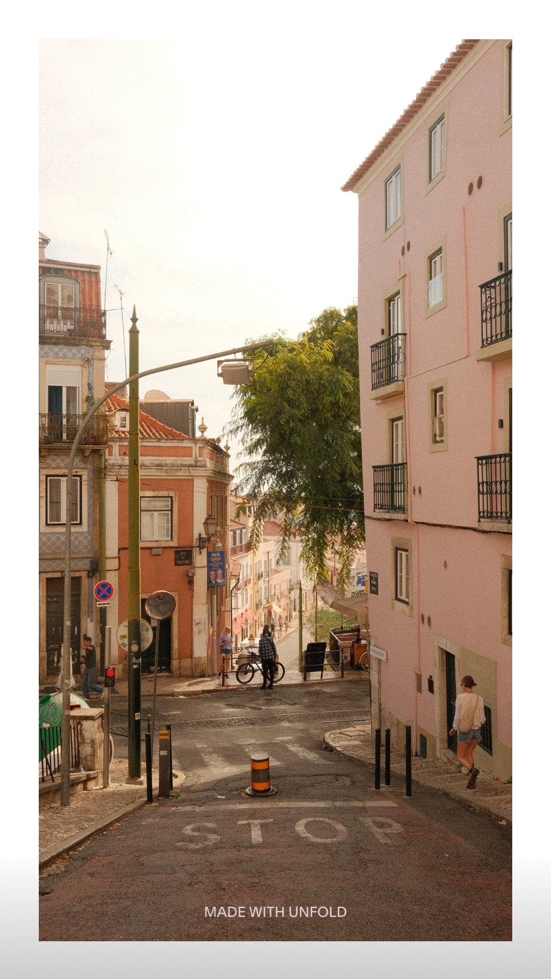 A narrow street in a European city with pastel-colored buildings, a stop sign painted on the road, and a few pedestrians and bicyclists at the intersection, with trees and a cloudy sky in the background.