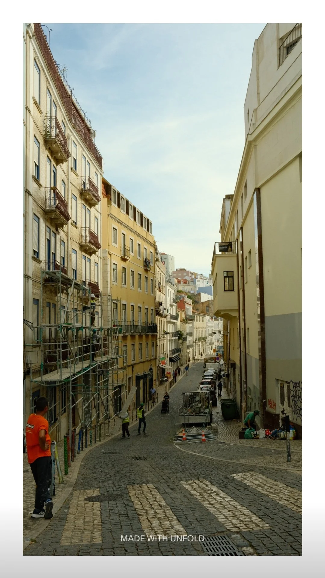 A narrow street in a city with multi-story buildings on both sides. Some buildings are under construction, with scaffolding on the left. People are walking and working along the street, which is paved with cobblestones. Cars are parked along the righ
