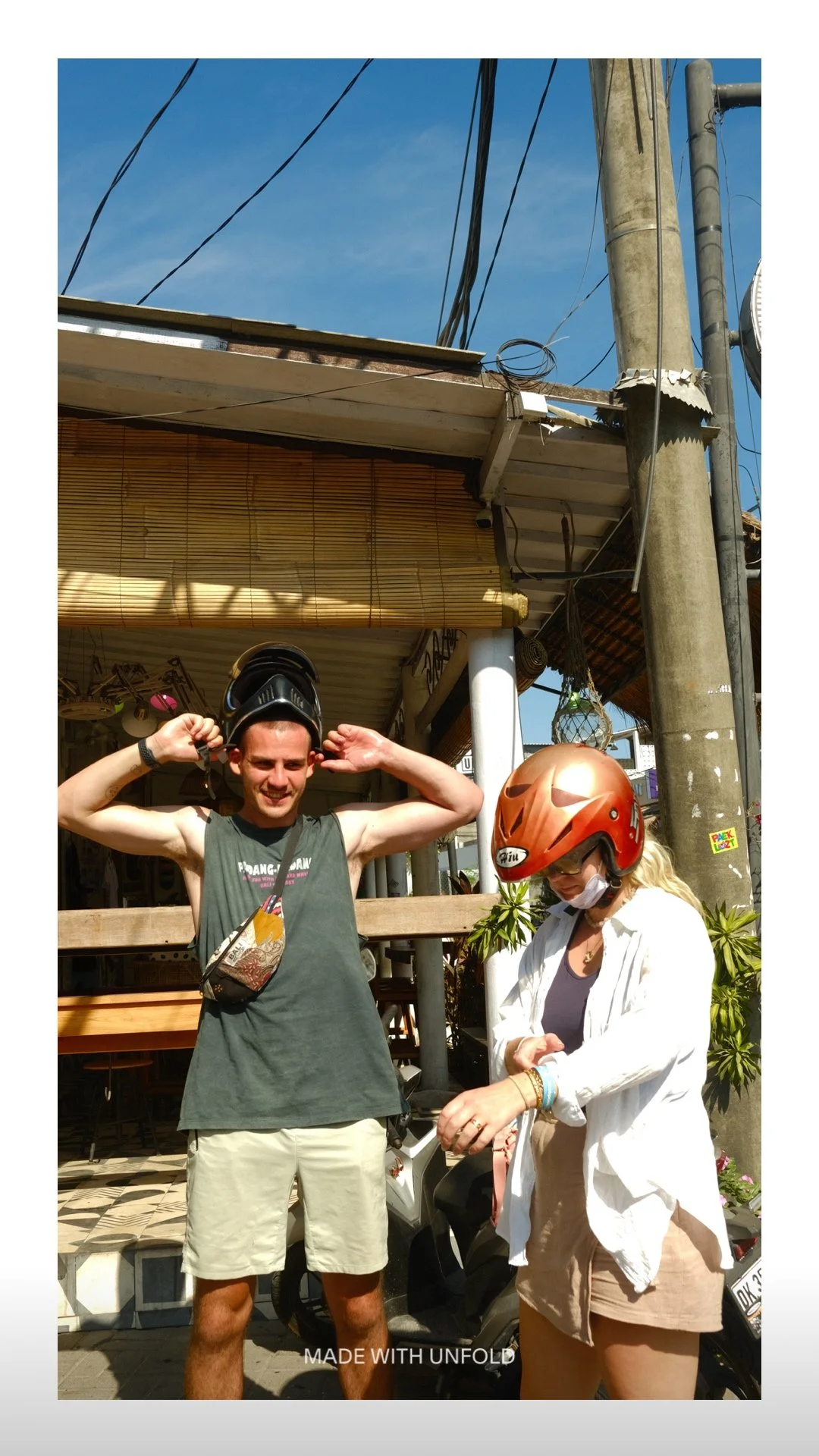 A young man and a woman wearing helmets stand outdoors under a clear blue sky. The young man is smiling and holds his helmet with both hands, while the woman, dressed in a white shirt and shorts, is adjusting her helmet. They are next to a motor scoo