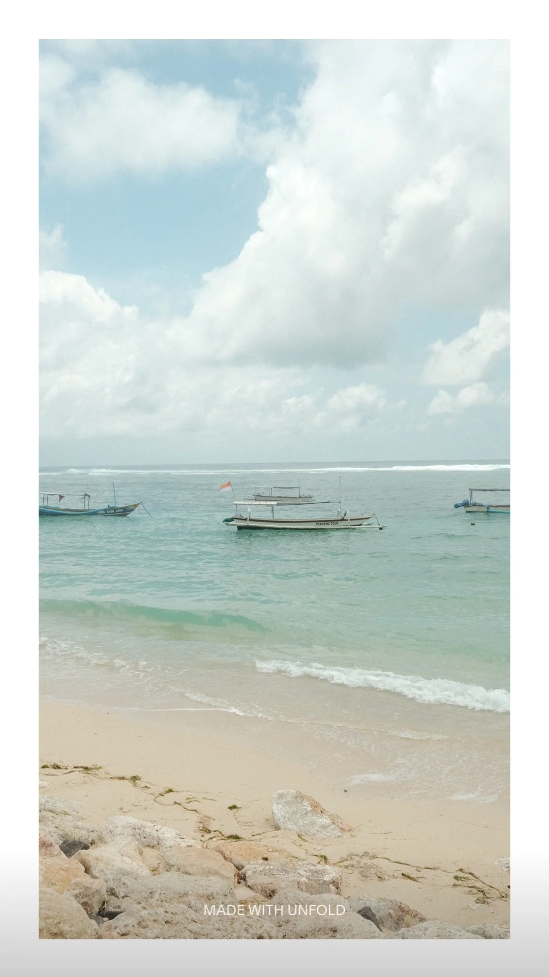 A beach with light sand, rocks, gentle waves, and several boats floating in the water under a partly cloudy sky.