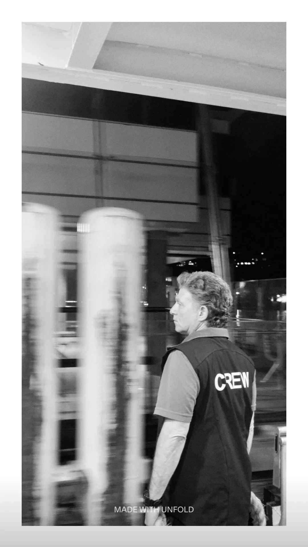 A black and white photo of a crew member with short curly hair, wearing a vest with "CREW" on the back, walking past blurred objects at night, possibly on an airport or industrial site.