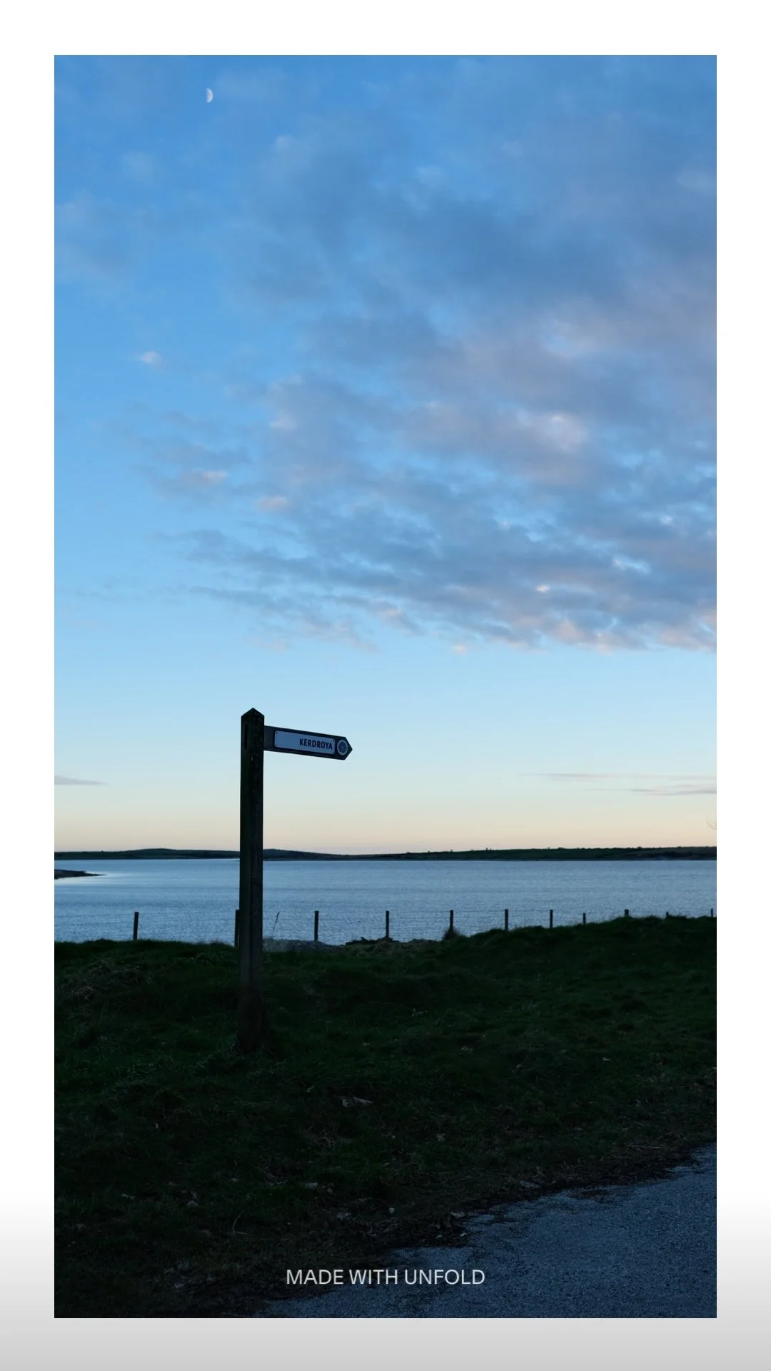 View of a lake or river with a signpost pointing to 'Kerodoya' under a partly cloudy sky in the evening.
