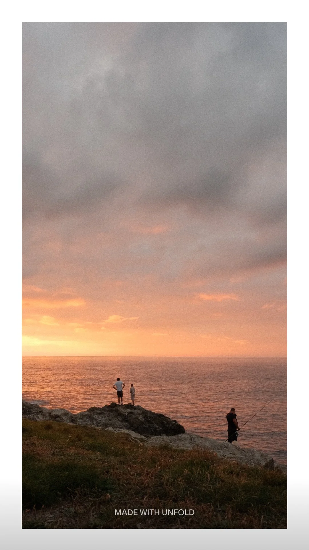 Three people on rocks at the edge of a body of water at sunset, two standing and one fishing, with a cloudy sky overhead.