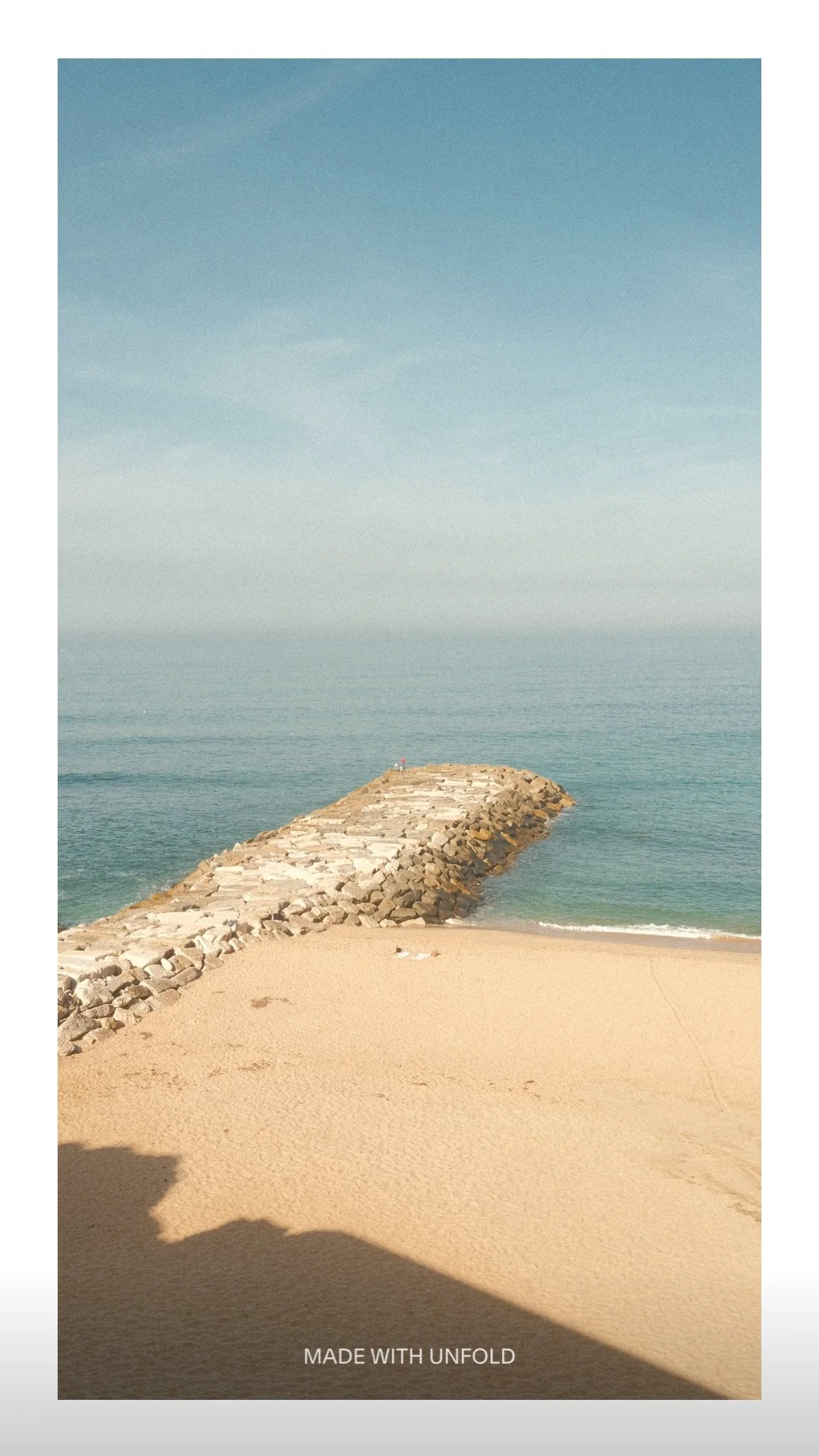 A sandy beach with a rocky breakwater extending into the ocean under a clear blue sky.