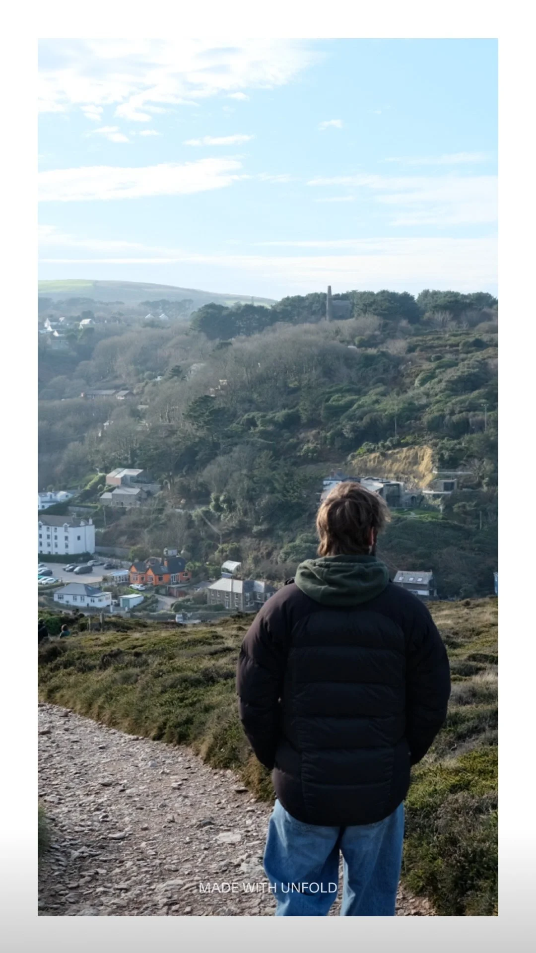 Man standing on a dirt path overlooking a hilly landscape with houses and trees, under a partly cloudy sky.