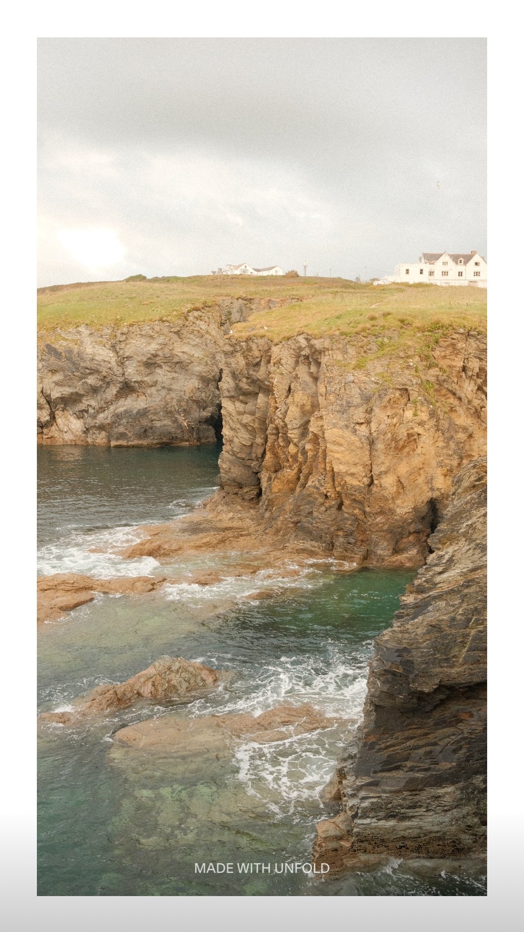 Cliffs and rocky shoreline by the ocean with houses on a grassy hill in the background under a cloudy sky.