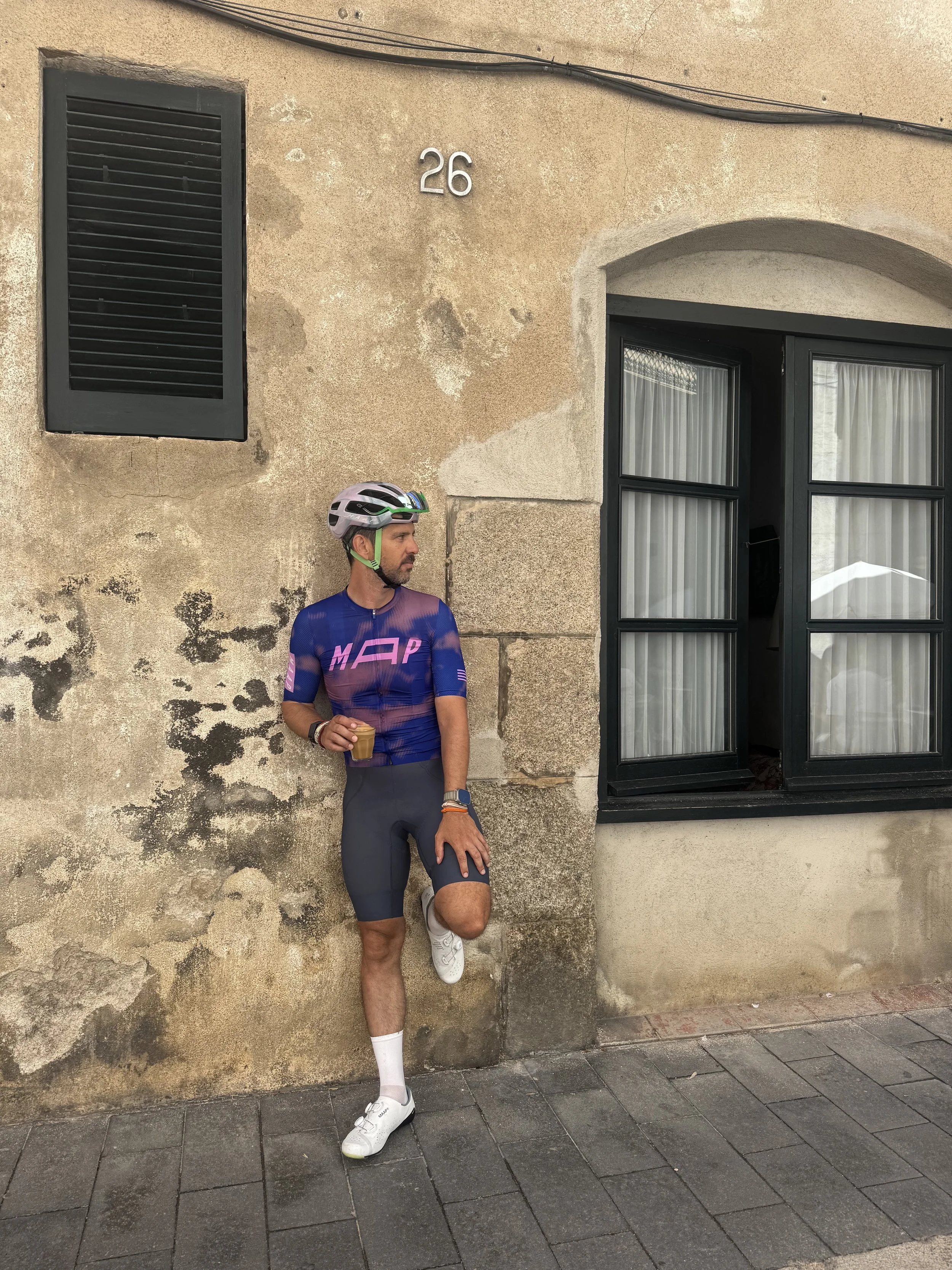 Hombre con casco y ropa de ciclismo, sosteniendo un vaso, apoyado en la pared de una casa antigua con ventana y número 26 en la pared.