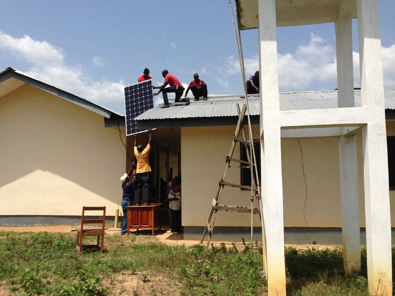 Man stands on a piece of furniture, passing a solar panel up to men on a metal roof in Africa