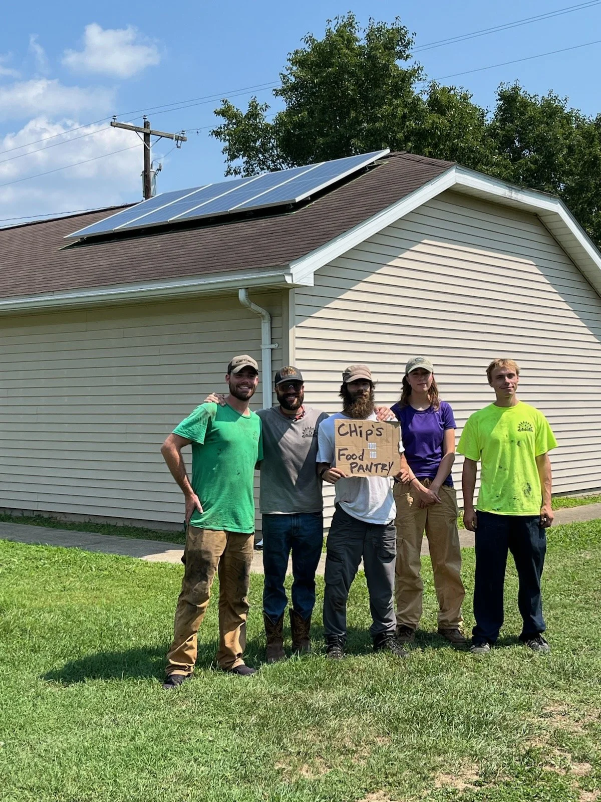 group of people holding "Chip's Food Pantry" sign, standing in front of a donated roof-mount solar system