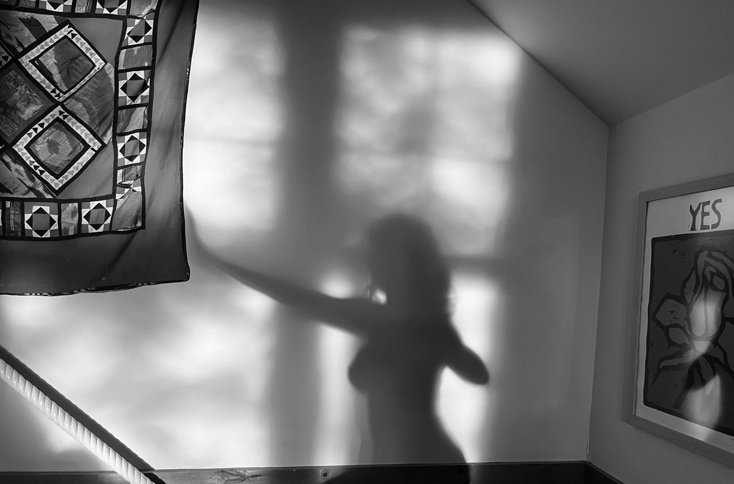 Black and white photograph showing a corner of a room with a quilted wall hanging, a shadow on the wall, and partially visible framed artwork with the word 'YES' on it.