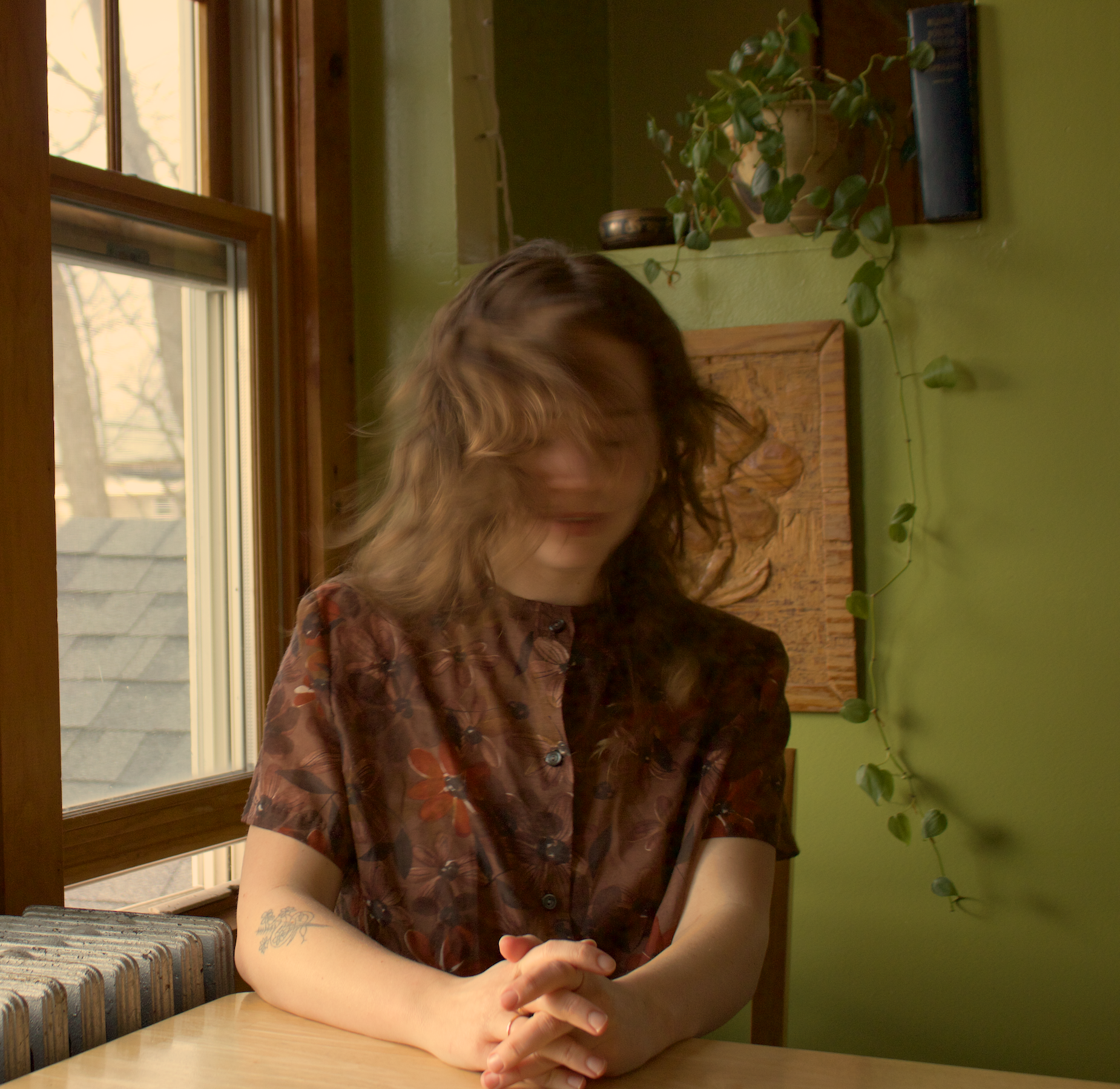 Woman with light brown wavy hair sitting at a wooden table, her arms crossed on the table, in front of a green wall with a string of leaf-shaped lights, near a large window, wearing a sheer brown floral dress.