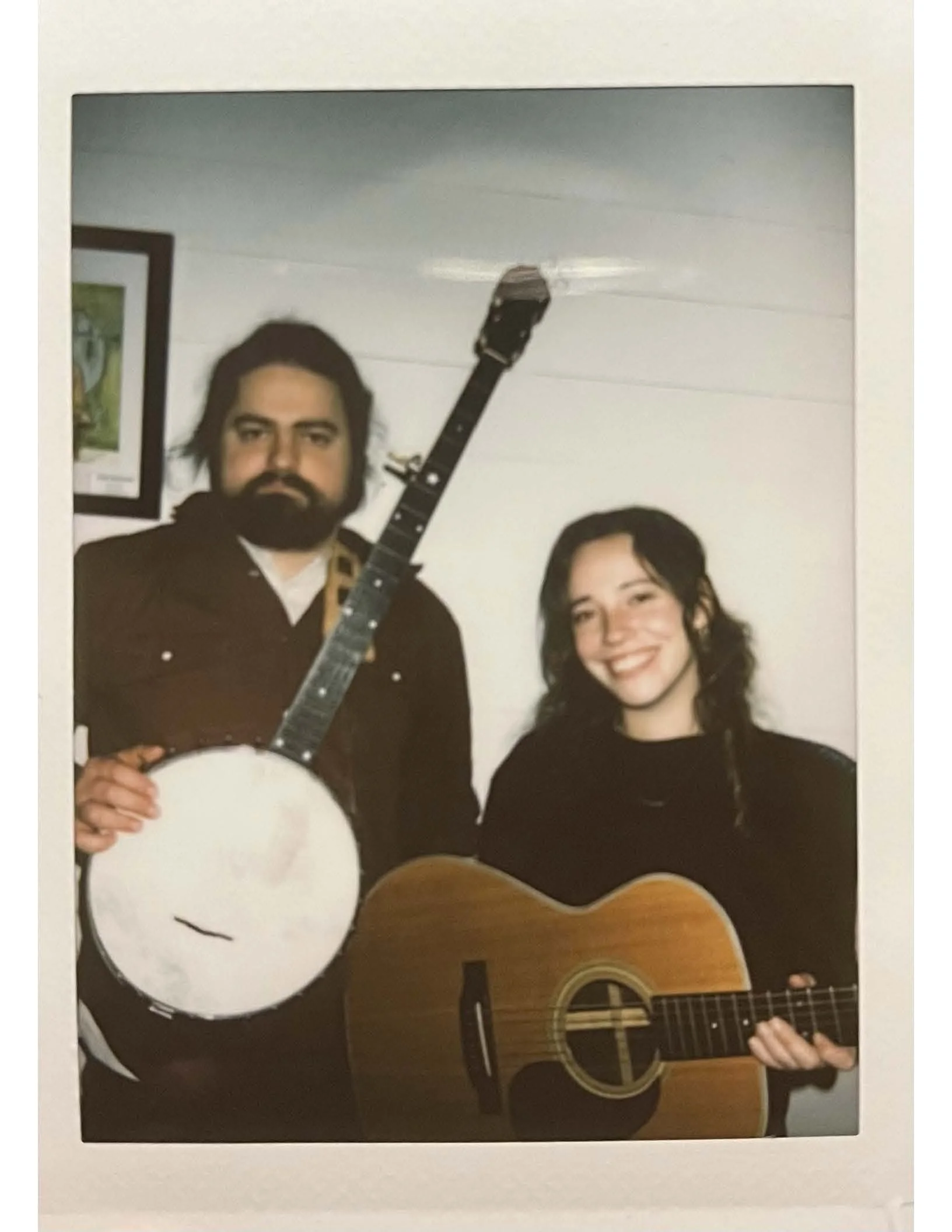 A framed photo shows a man with dark hair and a beard holding a banjo, standing next to a woman with long dark hair playing an acoustic guitar, both smiling. The background appears to be a plain white wall.