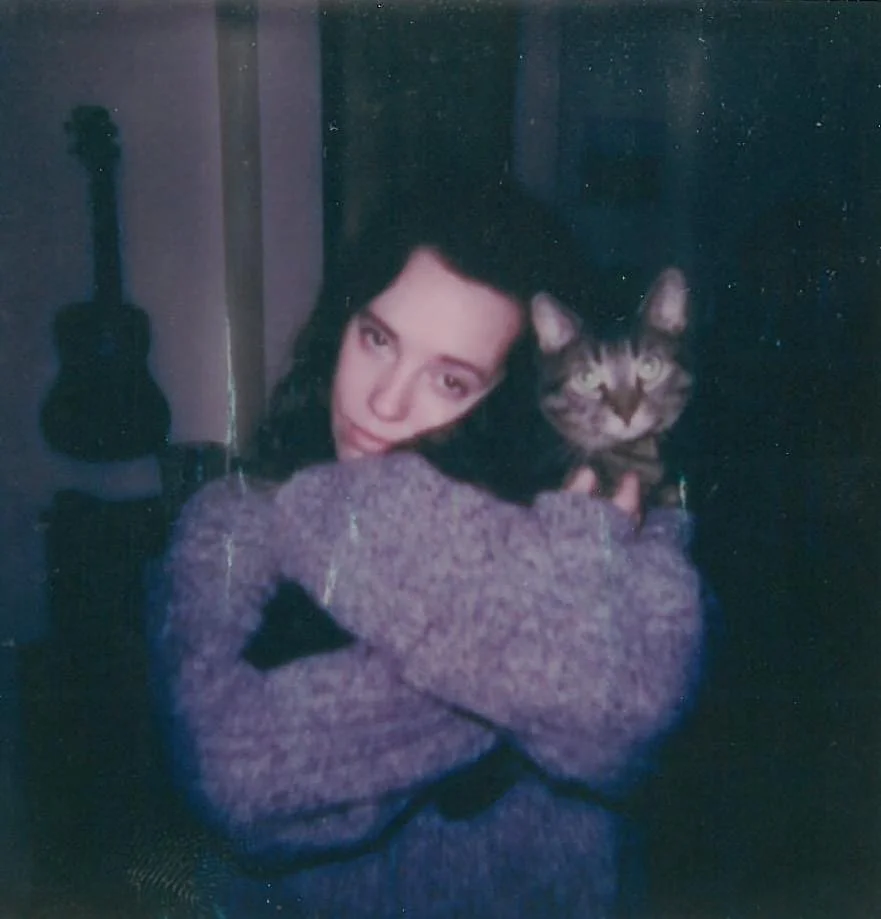 A woman with dark hair hugging a tabby cat, both looking at the camera, in a cozy indoor setting with a guitar in the background.