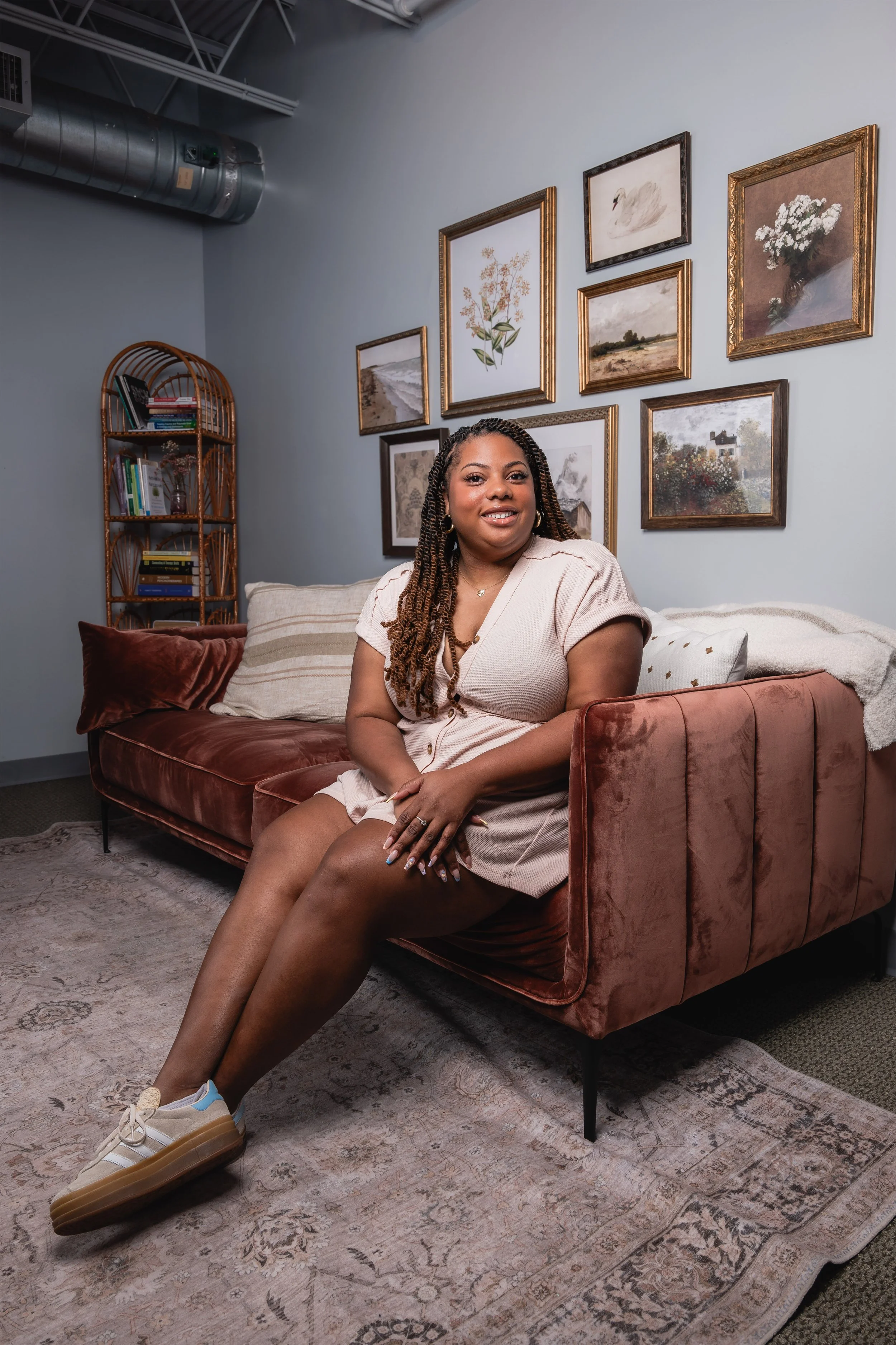 A woman sitting on a vintage velvet couch in a cozy living room, smiling at the camera. Behind her, there is a gallery wall with framed artworks, and a bookshelf in the corner.