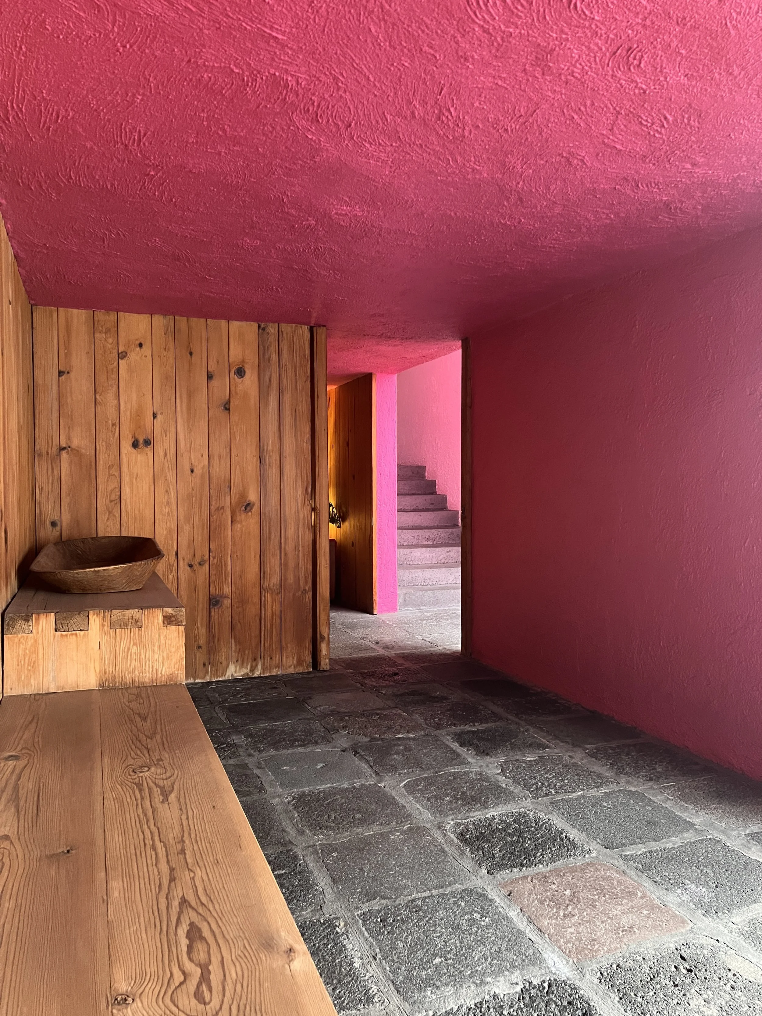 Interior of a room with pink and wooden walls, a pink ceiling, stone tile floor, a wooden bench, a wooden bowl, and a staircase in the background.