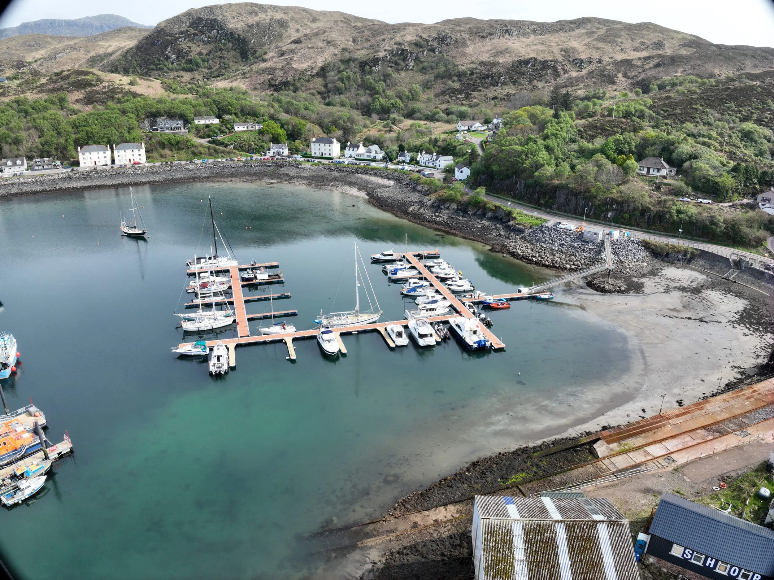 A harbor with moored boats, including a red fishing boat and a white boat, alongside a concrete pier with cars parked and buildings in the background.