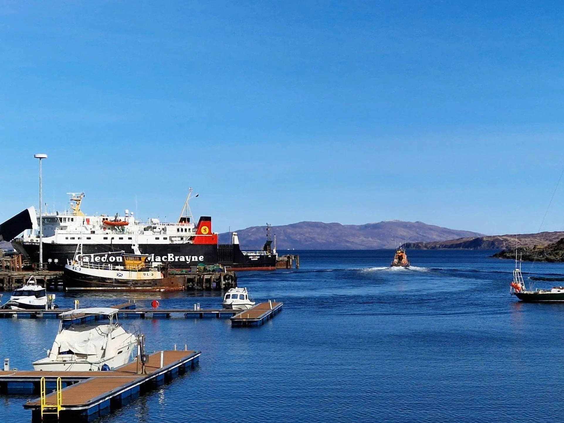 A harbor with sailboats and yachts docked along the pier, with colorful buildings and warehouses in the background under a clear blue sky.