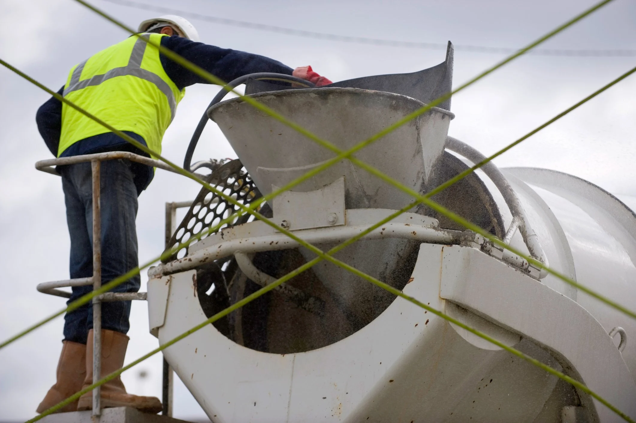 A construction worker in a yellow safety vest and brown boots operating a cement mixer, with green wires crossing in the foreground and an overcast sky in the background.