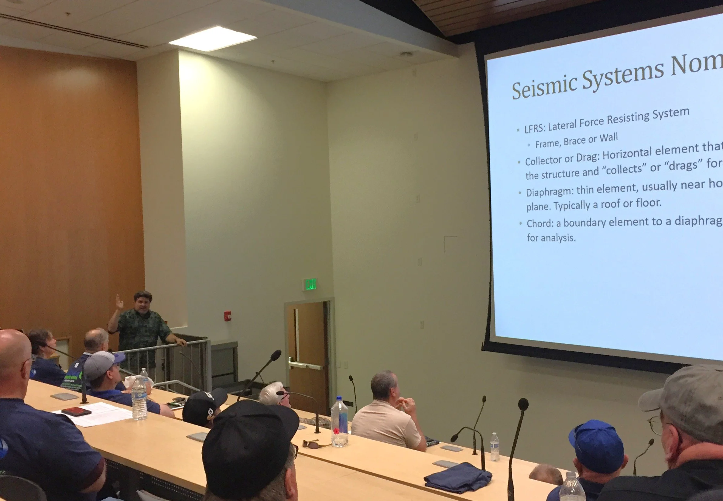 A man giving a presentation on seismic systems in a conference room with an audience. The presentation slide mentions LFRS, collector, diaphragm, and chord.